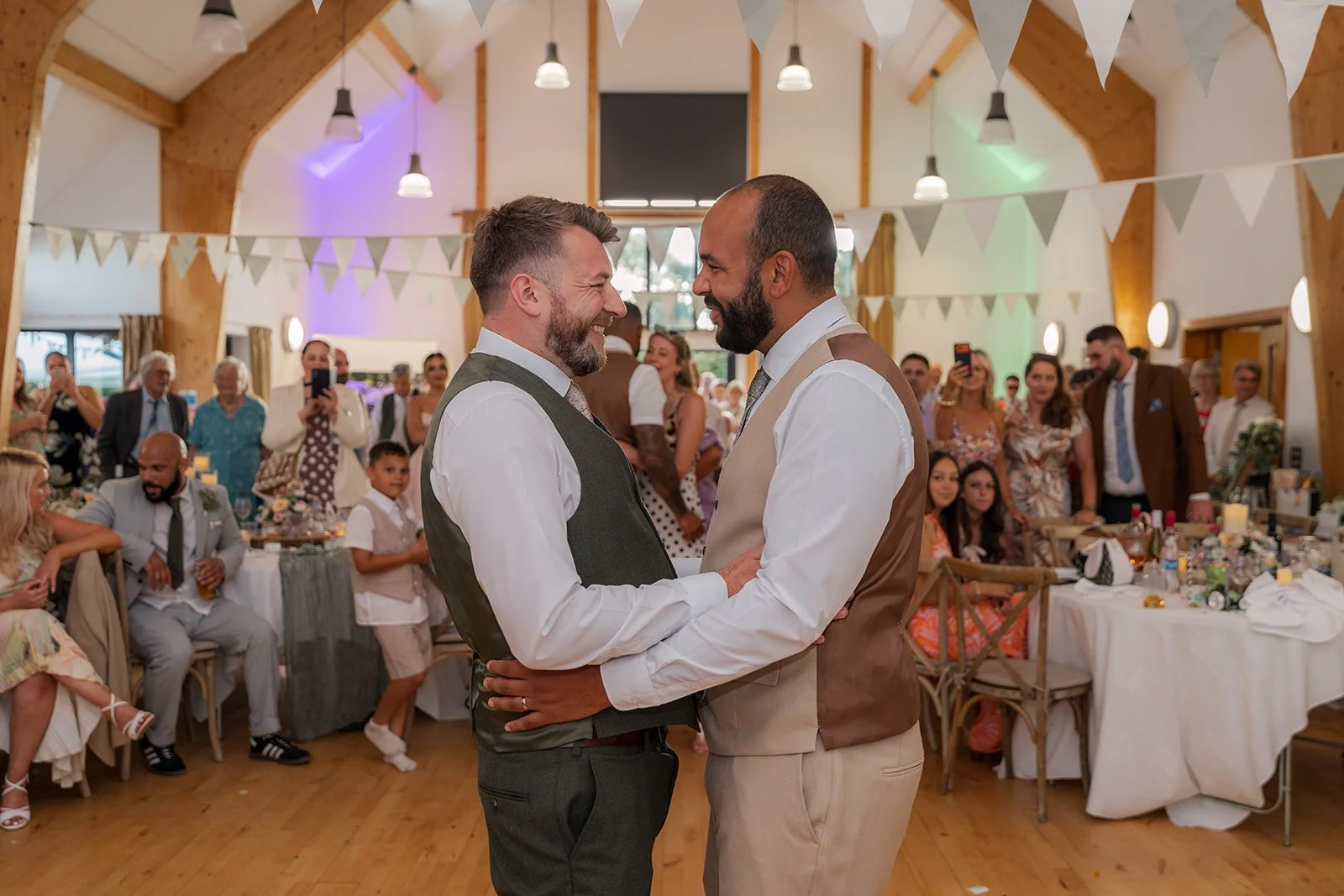 Aaron & Jack smile at one another during their first dance as wedding guests gather around them inside Rock Village Hall in Worcestershire, photographed by Just James Weddings.