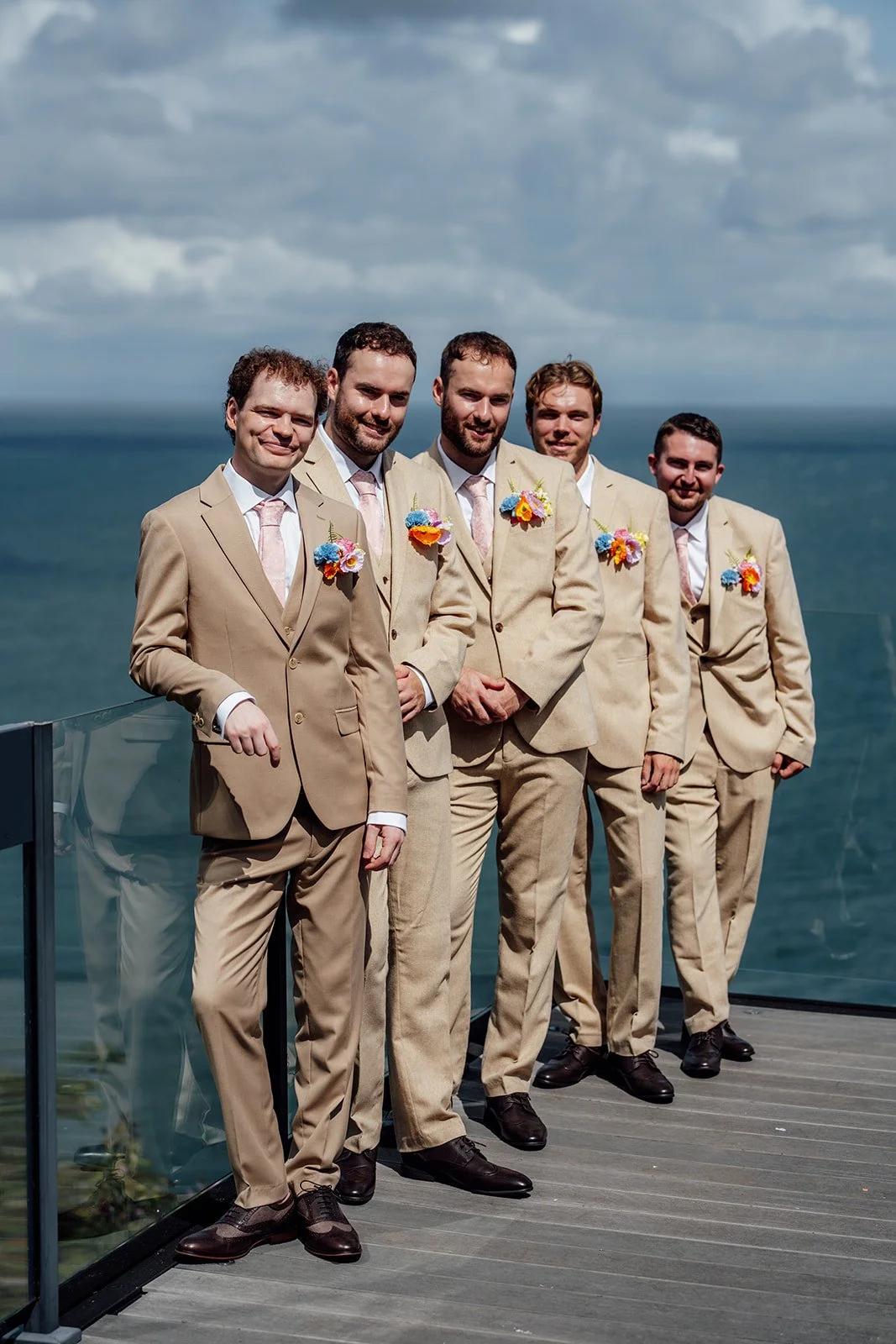 Alex poses with his groomsmen in matching beige suits and colourful buttonholes overlooking the sea at Sandy Cove Hotel in North Devon, photographed by Just James Weddings.