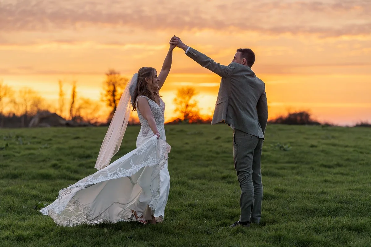 A bride and groom dancing outdoors during sunset, with the bride in a wedding dress and the groom in a suit.