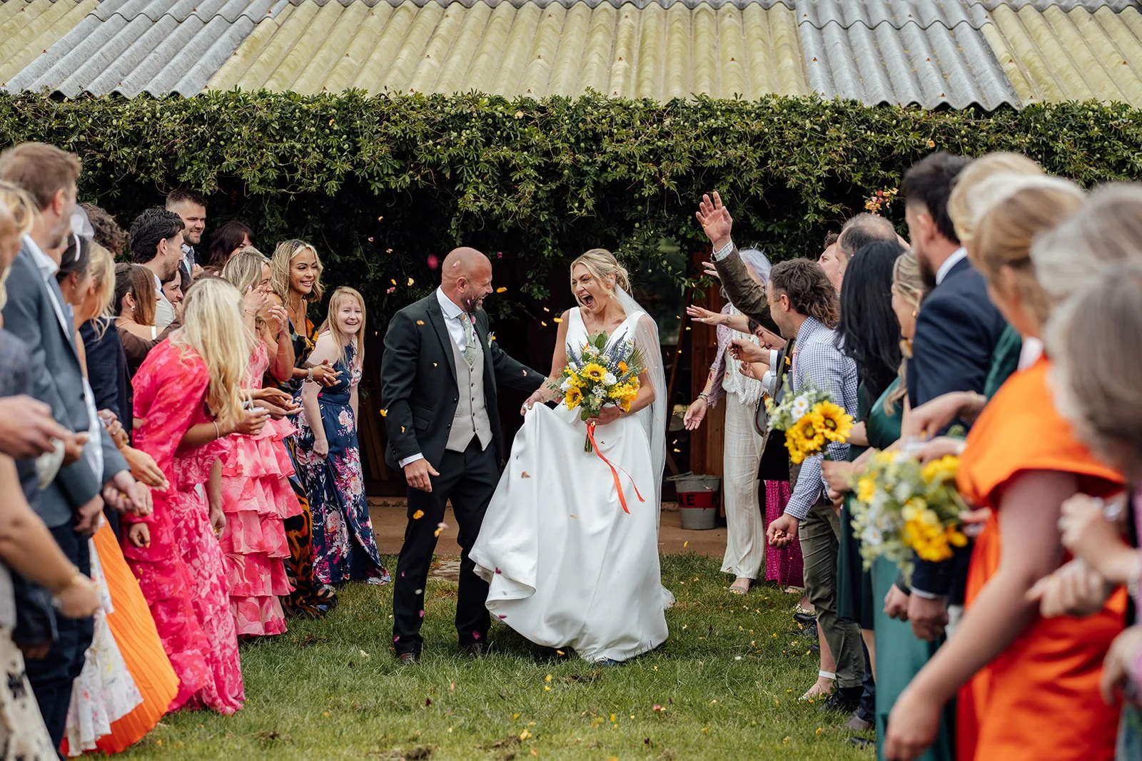 Colourful outdoor confetti photo of Hannah & Ben at Grays Barn in Devon, UK, laughing together as guests cheer and throw petals around them, with sunflowers and bold wedding outfits adding energy, photographed by Just James Weddings.