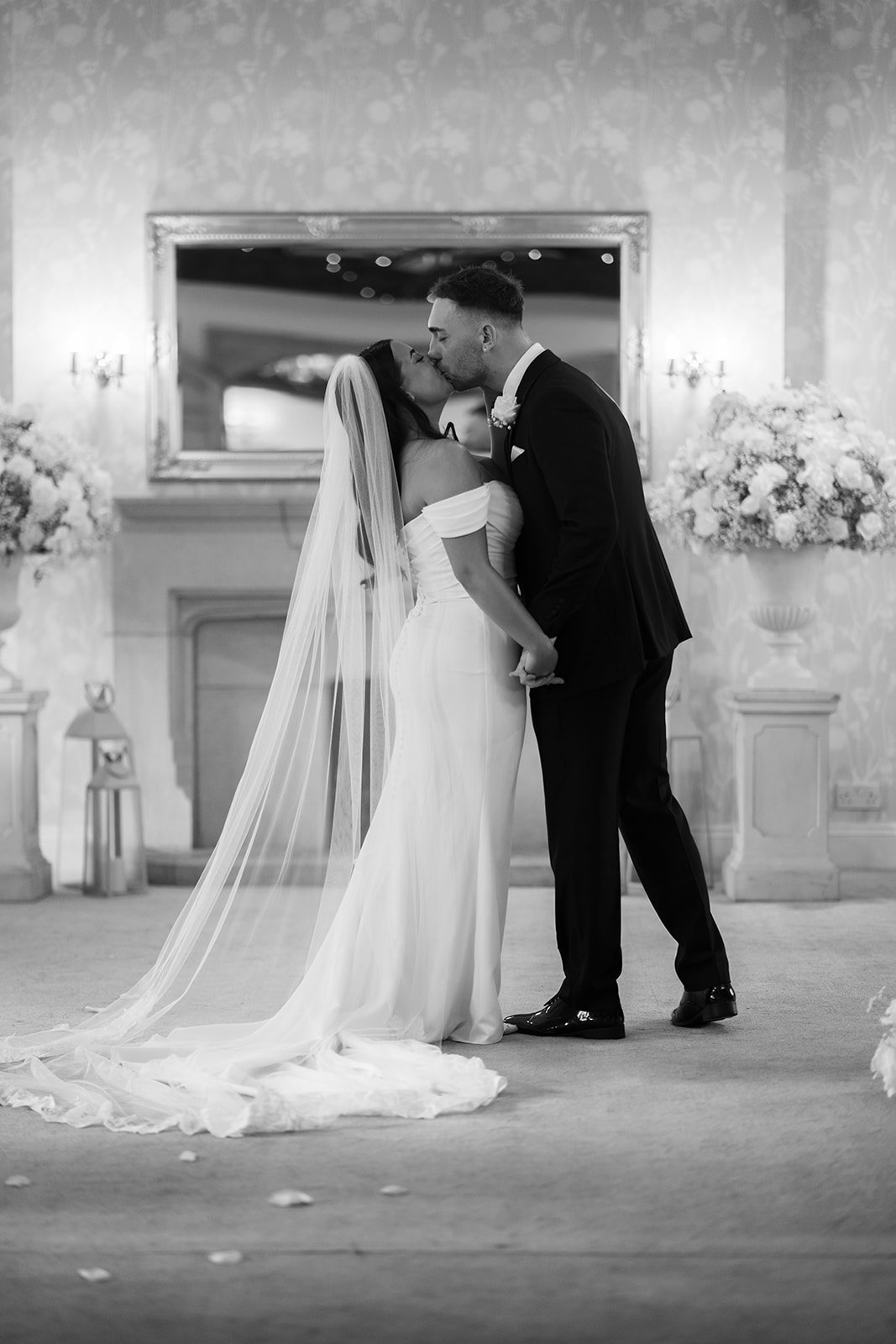 Black and white portrait of Nisha and Jack sharing a kiss inside Batch Country House in Somerset, UK, framed by a large mirror and white floral arrangements after their wedding ceremony during a quiet moment, photographed by Just James Weddings.