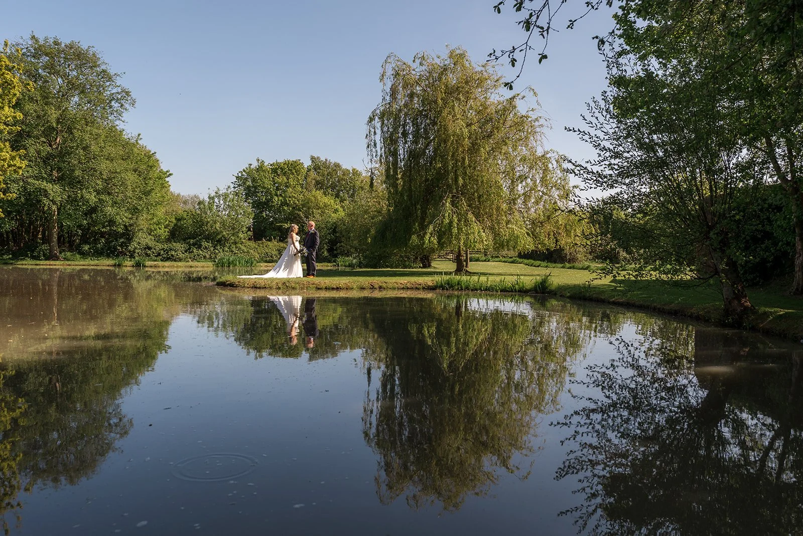 Debbie and Nigel share a peaceful lakeside portrait at Yarlington Barn in Somerset, standing beneath the trees as their reflection appears in the water for a calm countryside wedding.