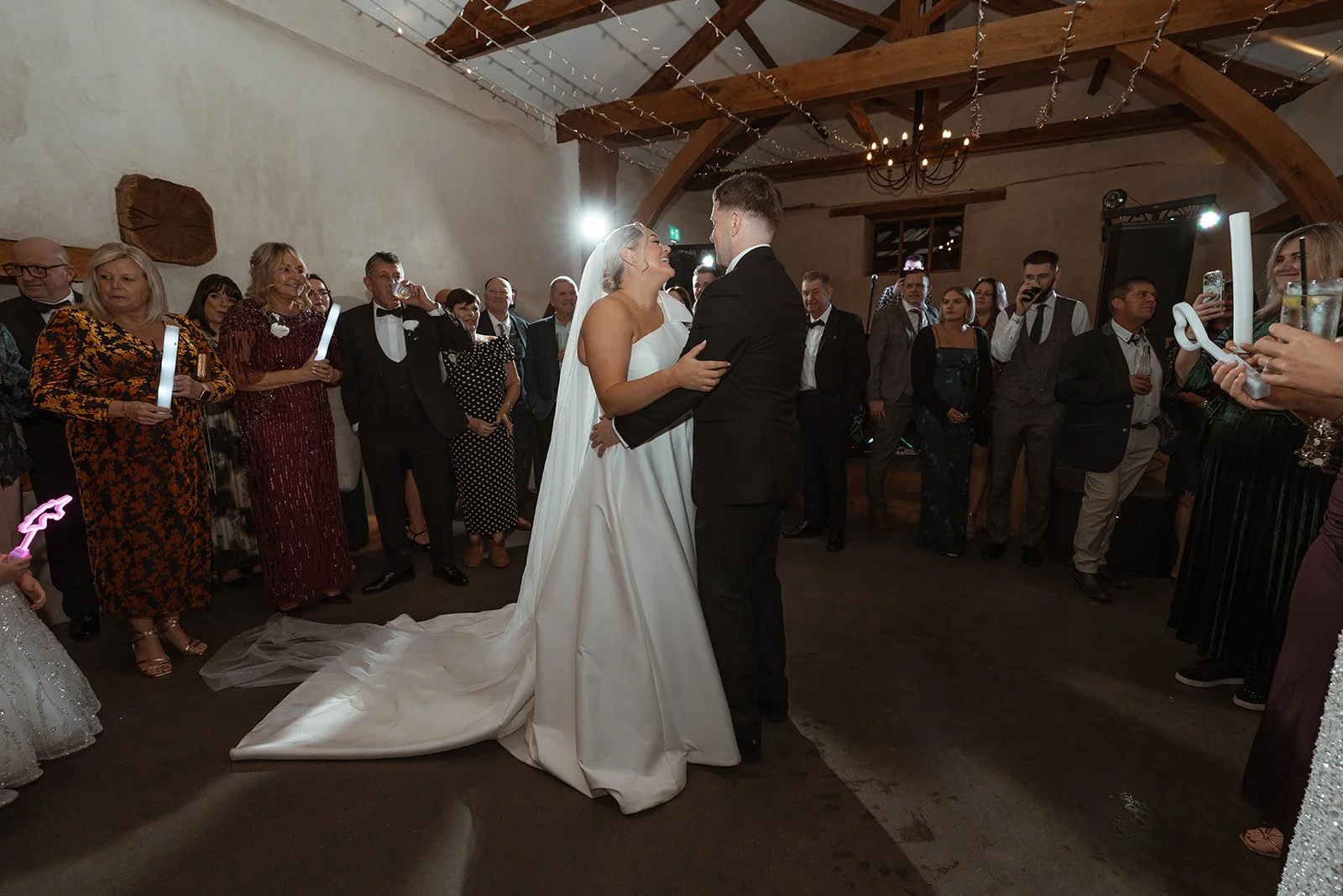 Jessie & Jay share their first dance surrounded by guests inside Upton Barn & Walled Garden in Devon, UK. Fairy lights and wooden beams glow overhead as friends and family watch, capturing joy, movement, and celebration on the dance floor.
