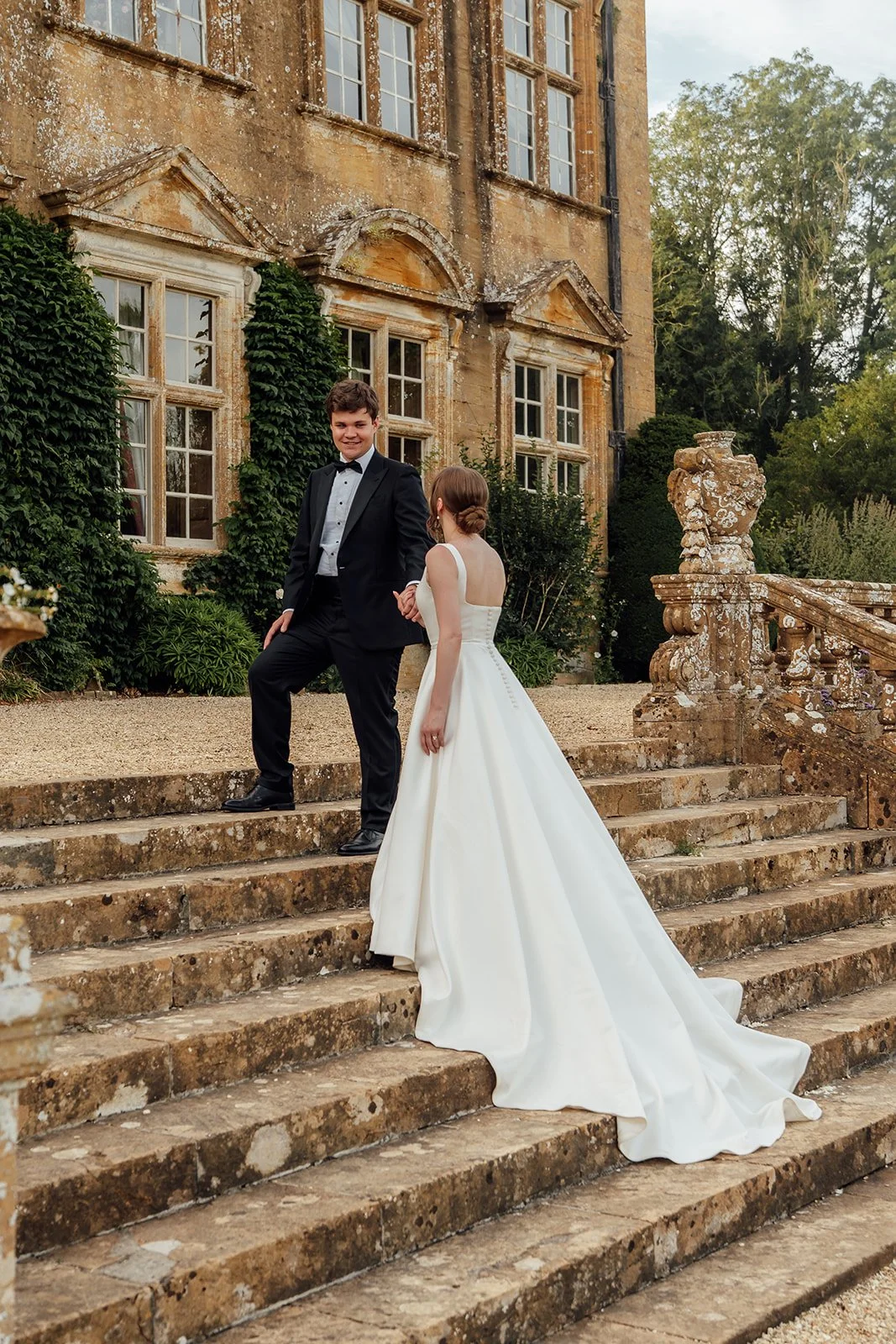 Rachel & Tom hold hands on the stone steps outside Brympton House, with the historic manor and gardens behind them in Somerset, photographed by Just James Weddings.