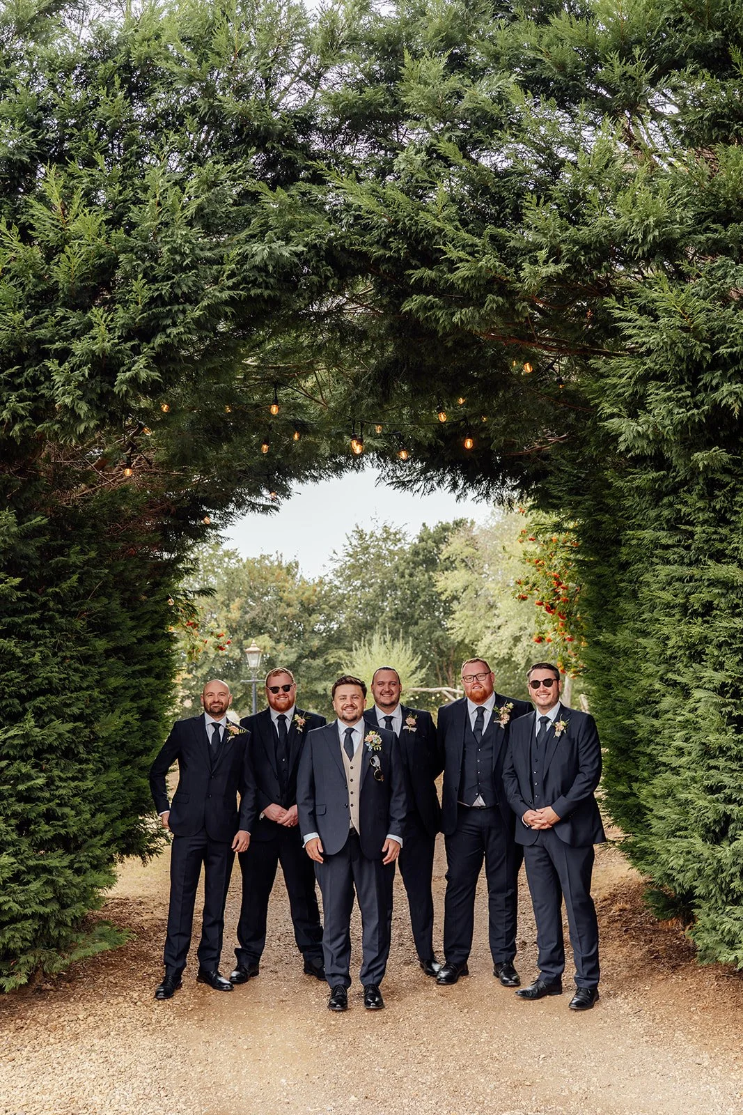 Simon poses with his groomsmen in navy suits beneath a leafy archway with festoon lights at Dovecote Events in Oxfordshire, photographed by Just James Weddings.