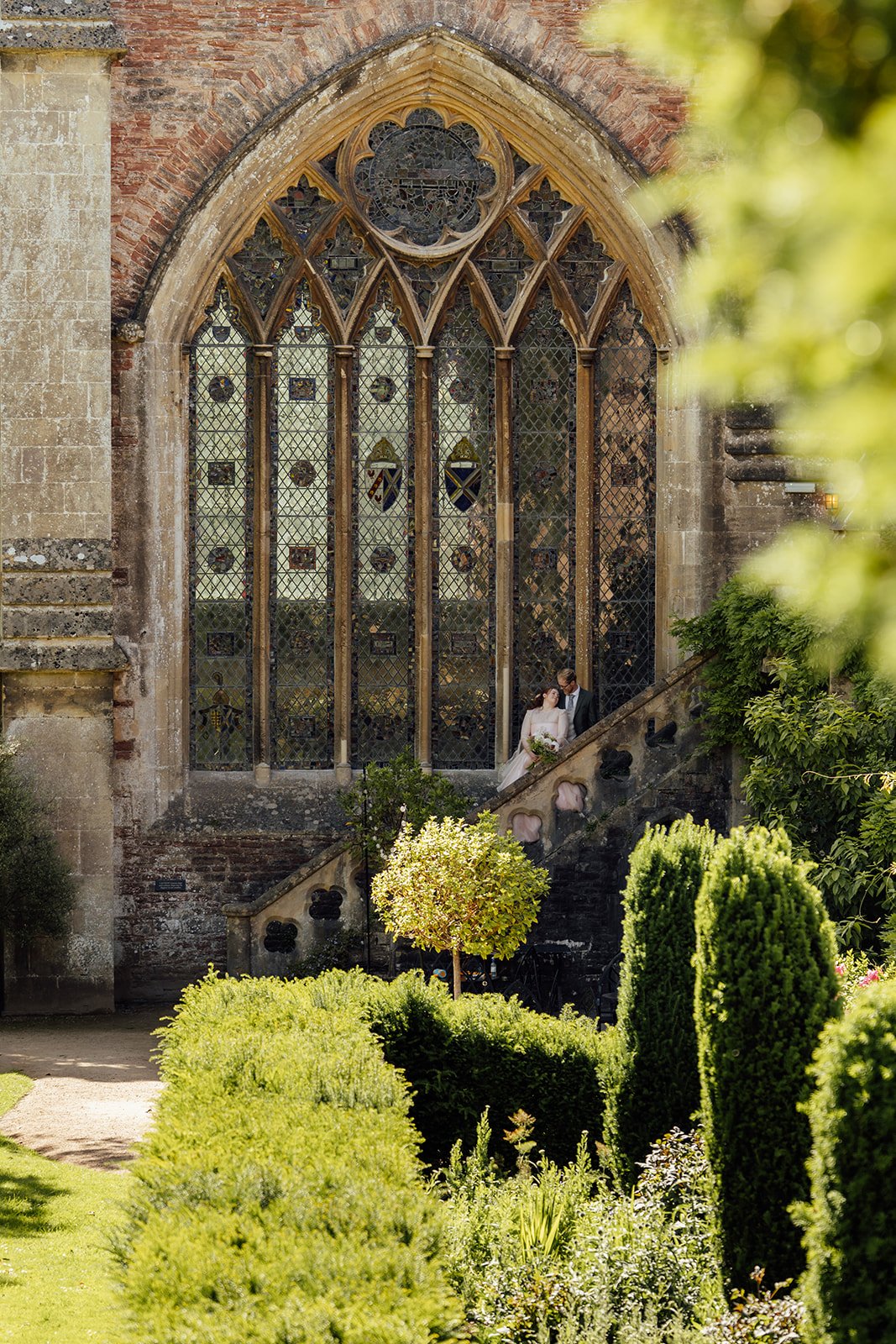 Charlotte & Jack couple portrait at Bishop’s Palace in Somerset, UK, showing them tucked on the stone steps beneath a huge stained glass window, framed by gardens and historic architecture during their sunny wedding day in Wells city centre.