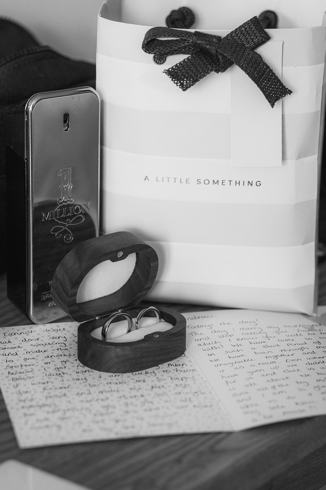 Groom’s wedding details from Andrew & Daisy’s Sandy Cove Hotel wedding in Devon, with wooden ring box holding wedding bands, handwritten letter, 1 Million fragrance and gift bag styled in black and white.