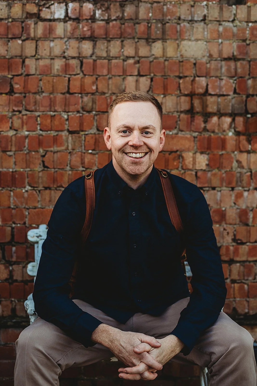 A smiling man with short light hair, wearing a black button-up shirt and tan pants, sitting on a white bench against a brick wall, with a leather backpack on his shoulders.