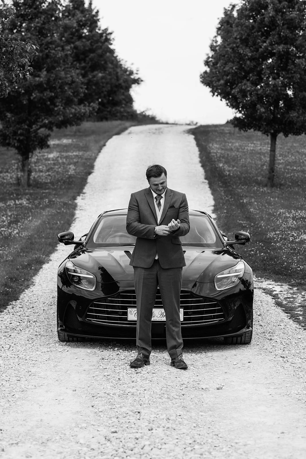 Armani adjusts his cuff while standing in front of a fast Aston Martin wedding car before marrying Chloe at The Barn at Upcote in Gloucestershire, photographed by Just James Weddings.
