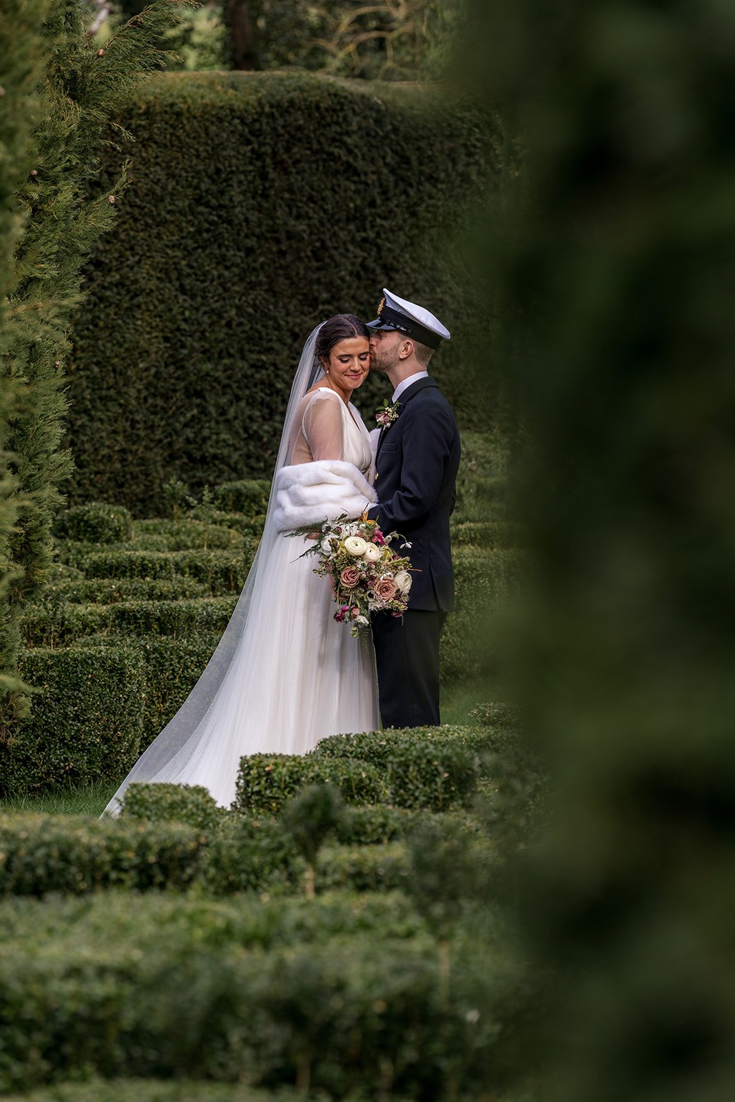 A bride and groom sharing a moment in a lush green garden, the bride in a white gown holding a bouquet, the groom in a navy suit with a sailor hat.