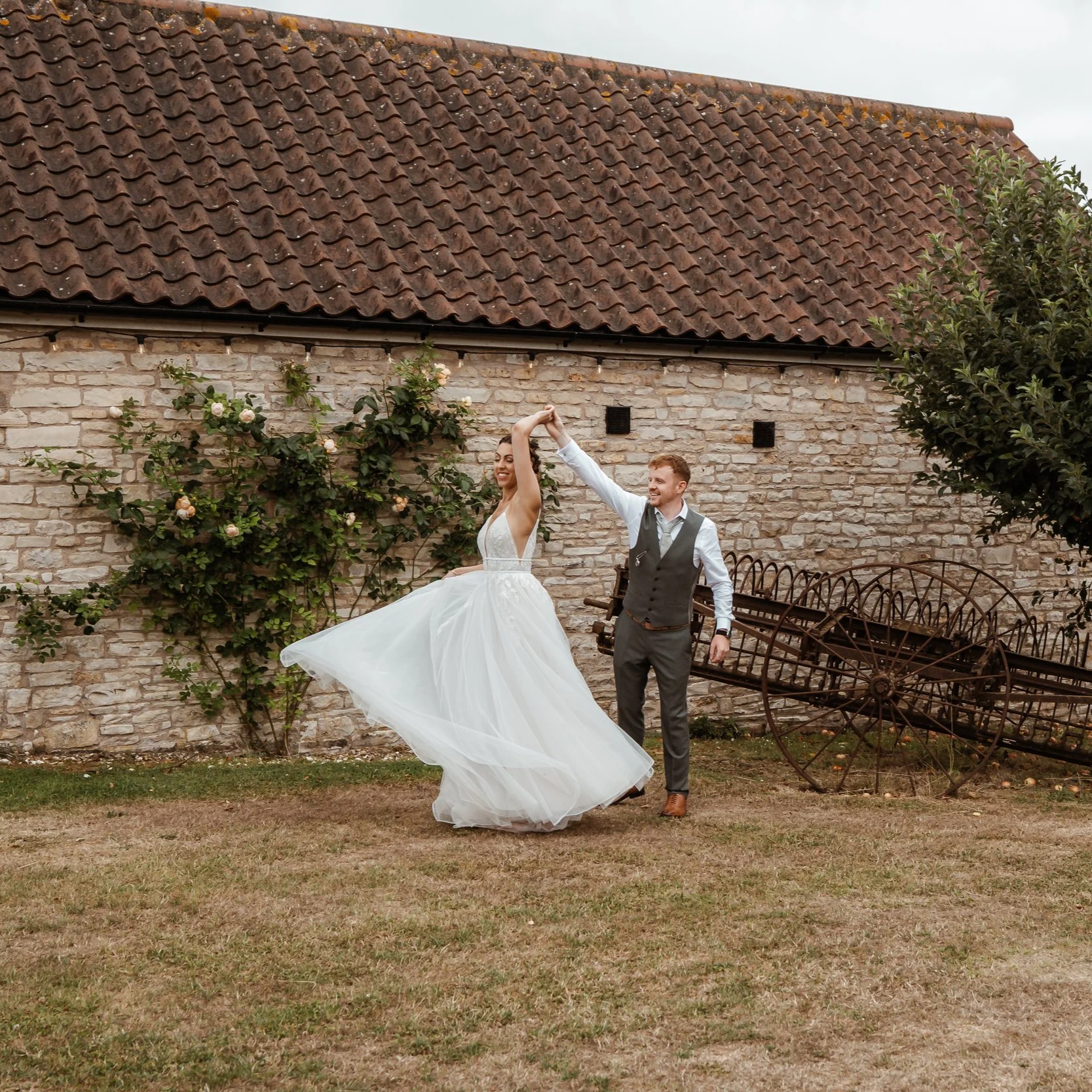 Madeleine & Izaac dancing together on the lawn at Folly Farm Centre, somerset, on their wedding day. The bride twirls in a flowing white gown while the groom lifts her hand, with a rustic stone wall, tiled roof, and vintage farm equipment behind.