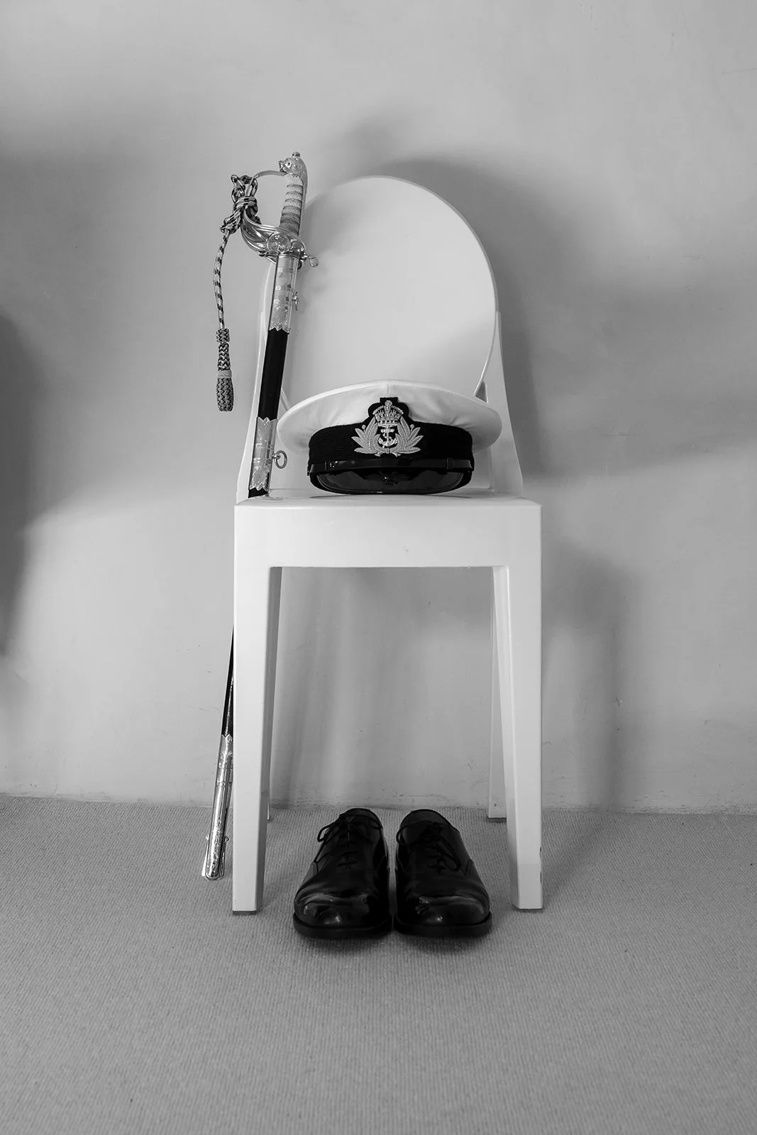 Wedding details for Megan & Luca at The Great Barn Devon show a naval dress sword, white peaked cap and polished black shoes carefully arranged on a white chair against a simple wall, captured in timeless black and white inside the Devon venue.