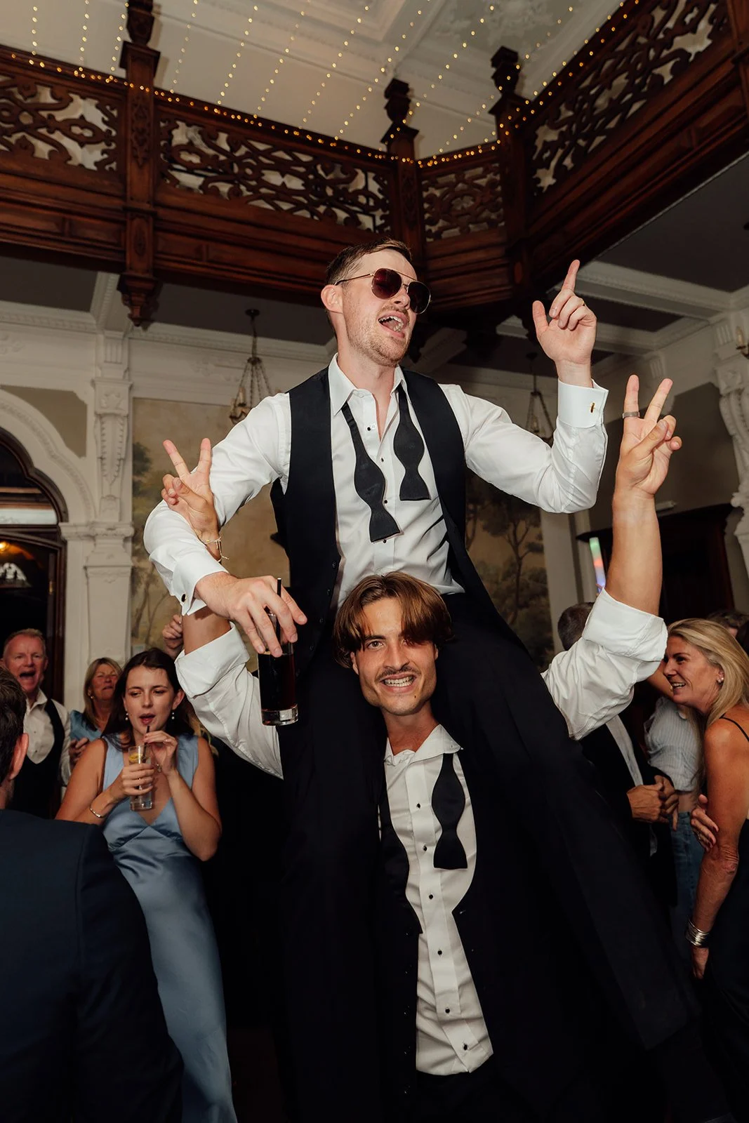 Wedding guests celebrating on the dance floor at Caitlin and Kevin’s Clevedon Hall wedding in Somerset, with one guest lifted onto another’s shoulders beneath the venue’s ornate interior.