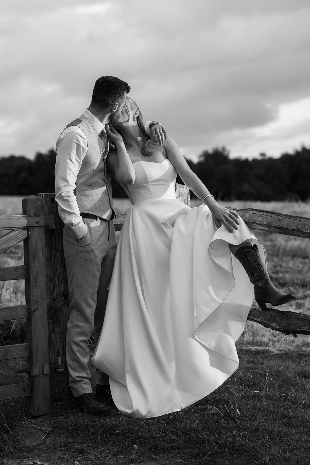 Black and white portrait of Louise & Christian kissing on a wooden field gate at The Cider Barn in Somerset, photographed by Just James Weddings for romantic countryside wedding photography.