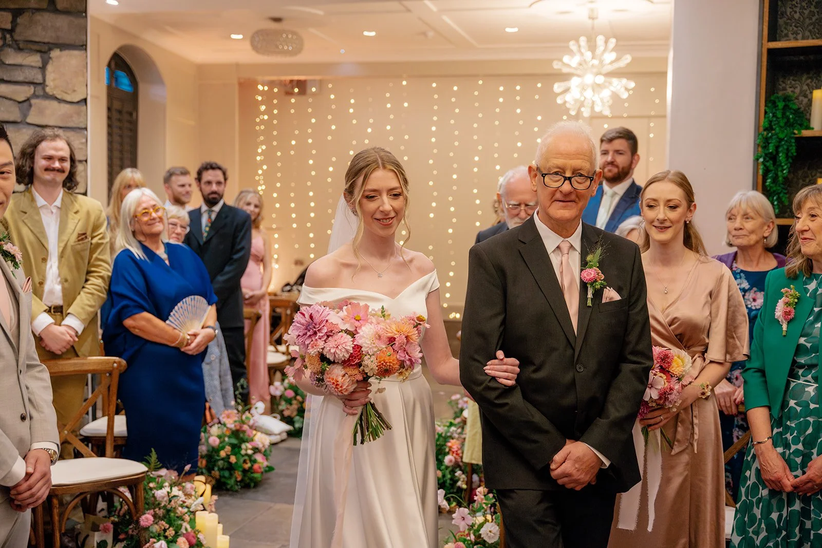 Josie walking down the aisle with her father at Clifton Observatory in Somerset, holding a pink bouquet and smiling at guests, photographed by Just James Weddings.