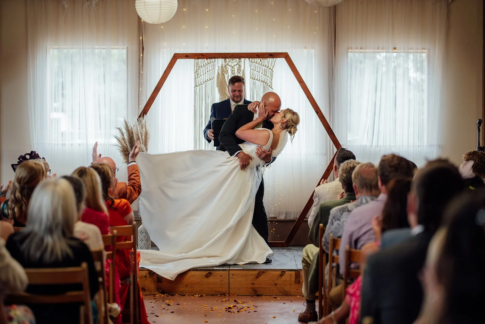Joyful wedding ceremony moment as Hannah & Ben share a dramatic first kiss at Grays Barn in Devon, UK, surrounded by cheering guests, soft draping, fairy lights and a wooden arch backdrop, photographed with energy by Just James Weddings.