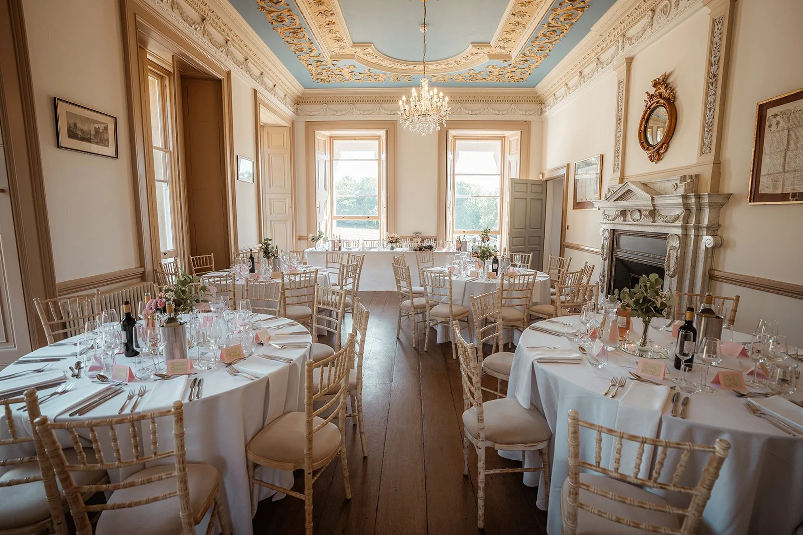 Wedding reception room at Crowcombe Court in Somerset, UK, set with elegant round tables, glassware and chairs beneath an ornate ceiling for Michelle and Andy’s celebration.