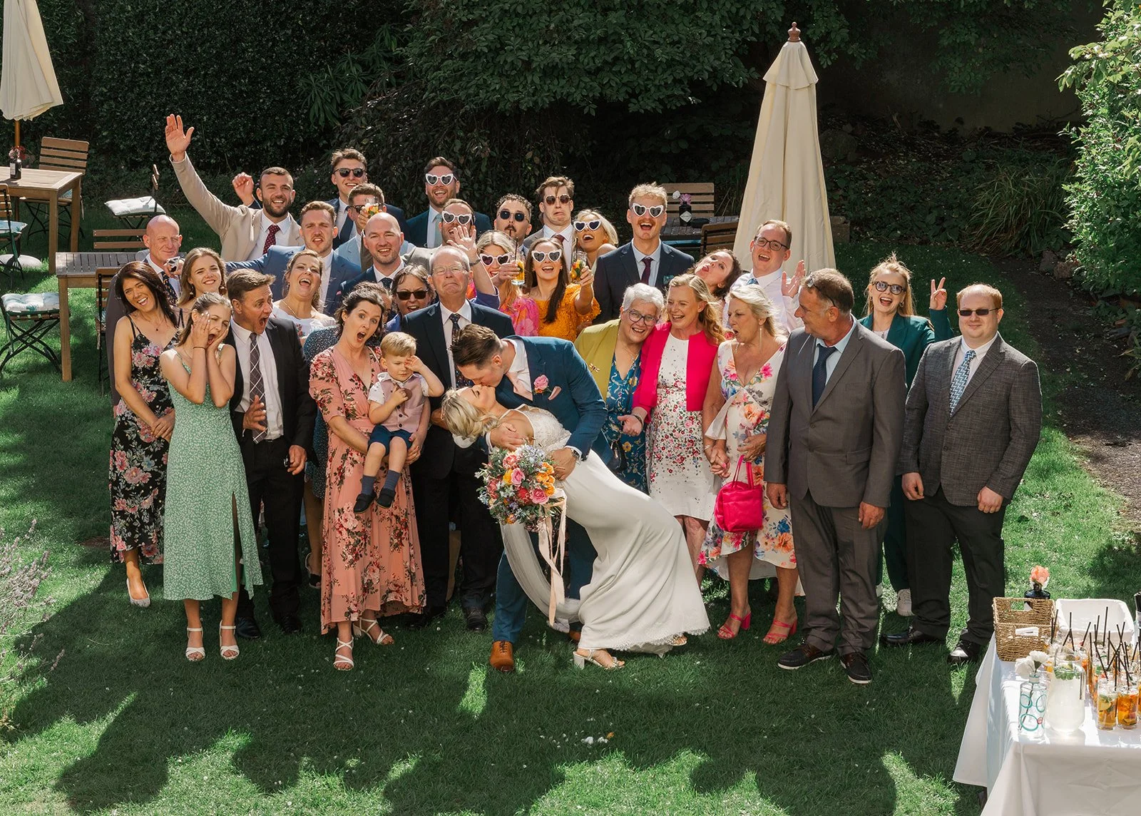 Kristy and Will celebrate with friends and family during a fun outdoor group wedding photo at No. 4 Clifton Village in Bristol, UK, with guests cheering around the newlyweds.