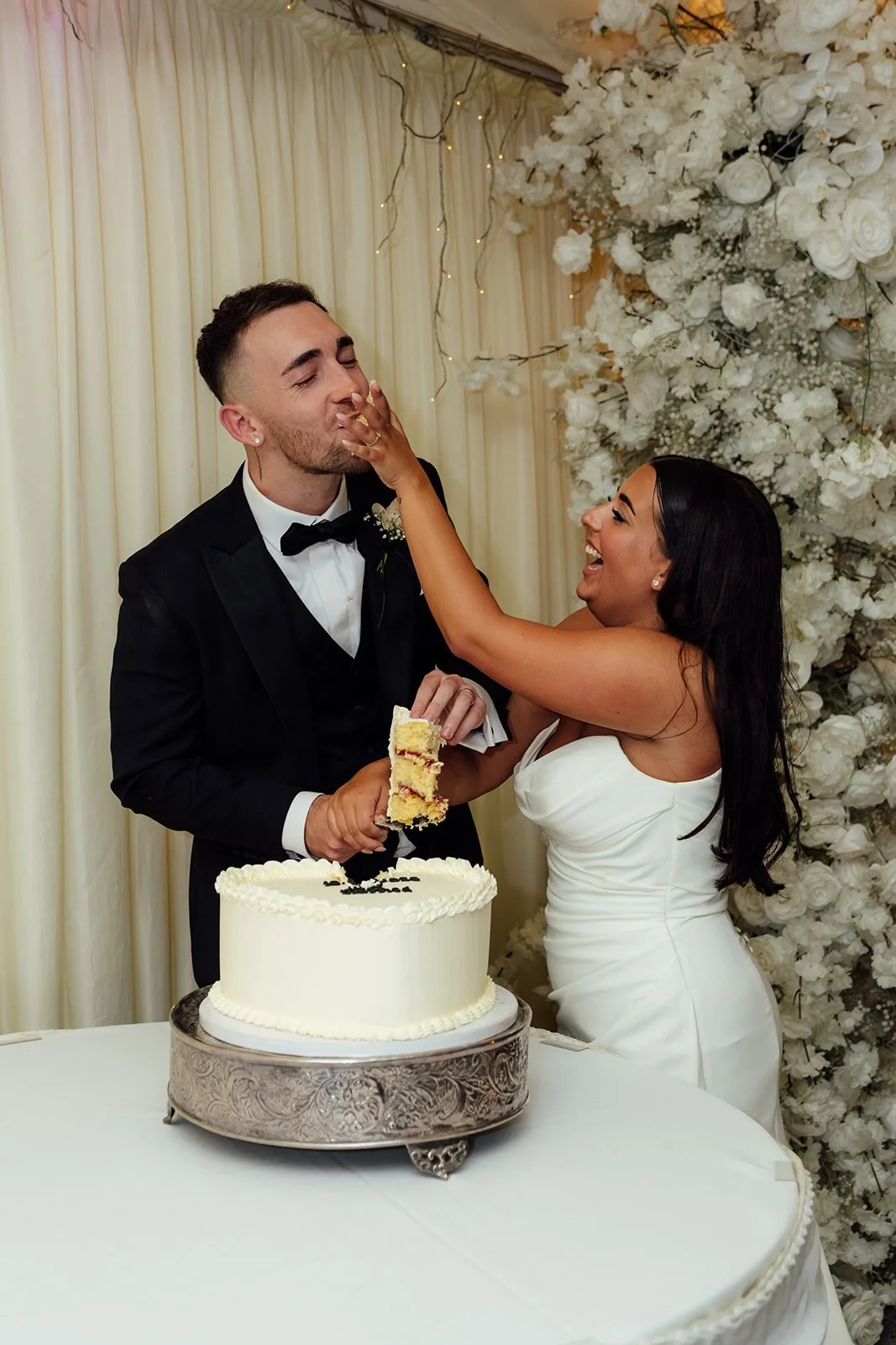 Nisha laughs as she feeds Jack wedding cake during their reception at Batch Country House in Somerset, UK, with a white floral backdrop and fairy lights behind them, capturing a playful newlywed moment photographed by Just James Weddings.