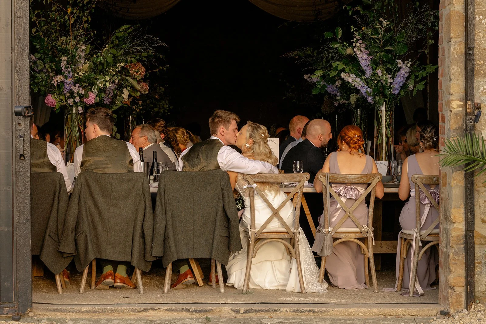 Abi & Josh sitting together during their wedding breakfast at The Cider Barn in Somerset, surrounded by guests, flowers and stone barn details, photographed by Just James Weddings for relaxed reception moments.