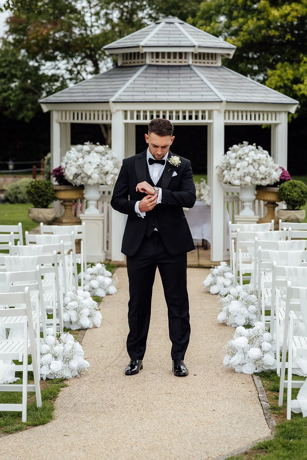 Jack adjusts his cufflinks in a black tuxedo beside the white garden gazebo at Batch Country House in Somerset, UK, surrounded by white floral arrangements and ceremony chairs before marrying Nisha there, photographed by Just James Weddings.