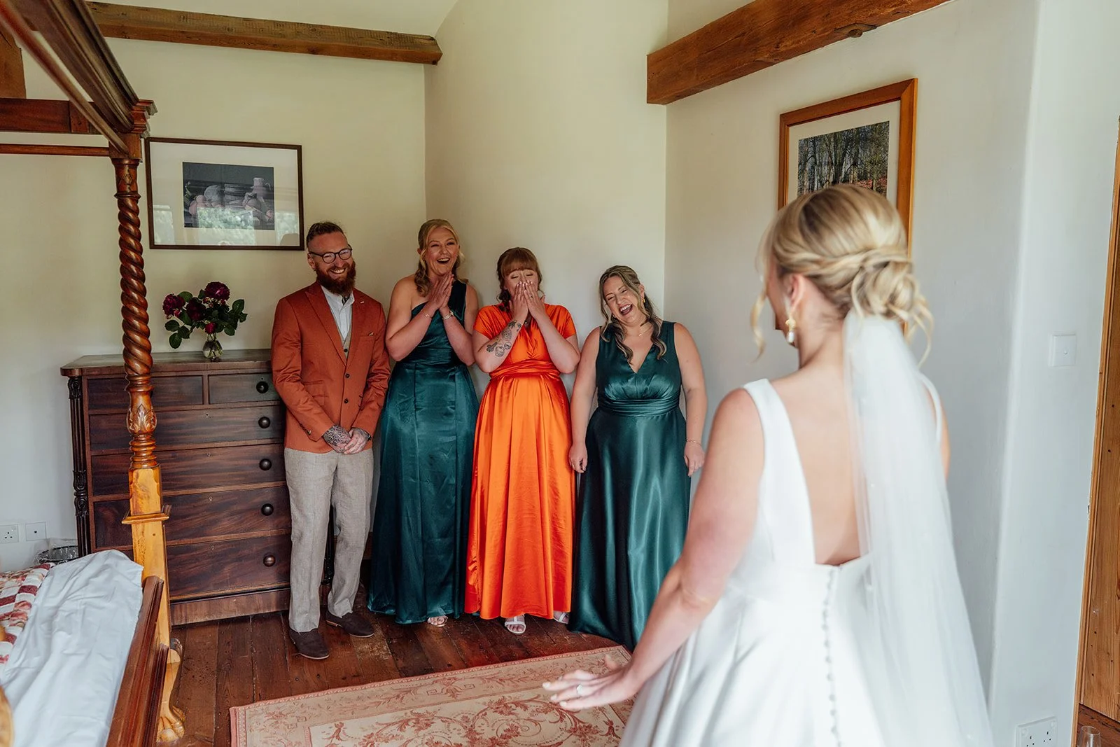 Emotional first look with Hannah’s wedding party at Grays Barn in Devon, UK, as friends react with laughter and happy tears to the bride in her gown and veil inside the rustic bedroom, photographed naturally and warmly by Just James Weddings.