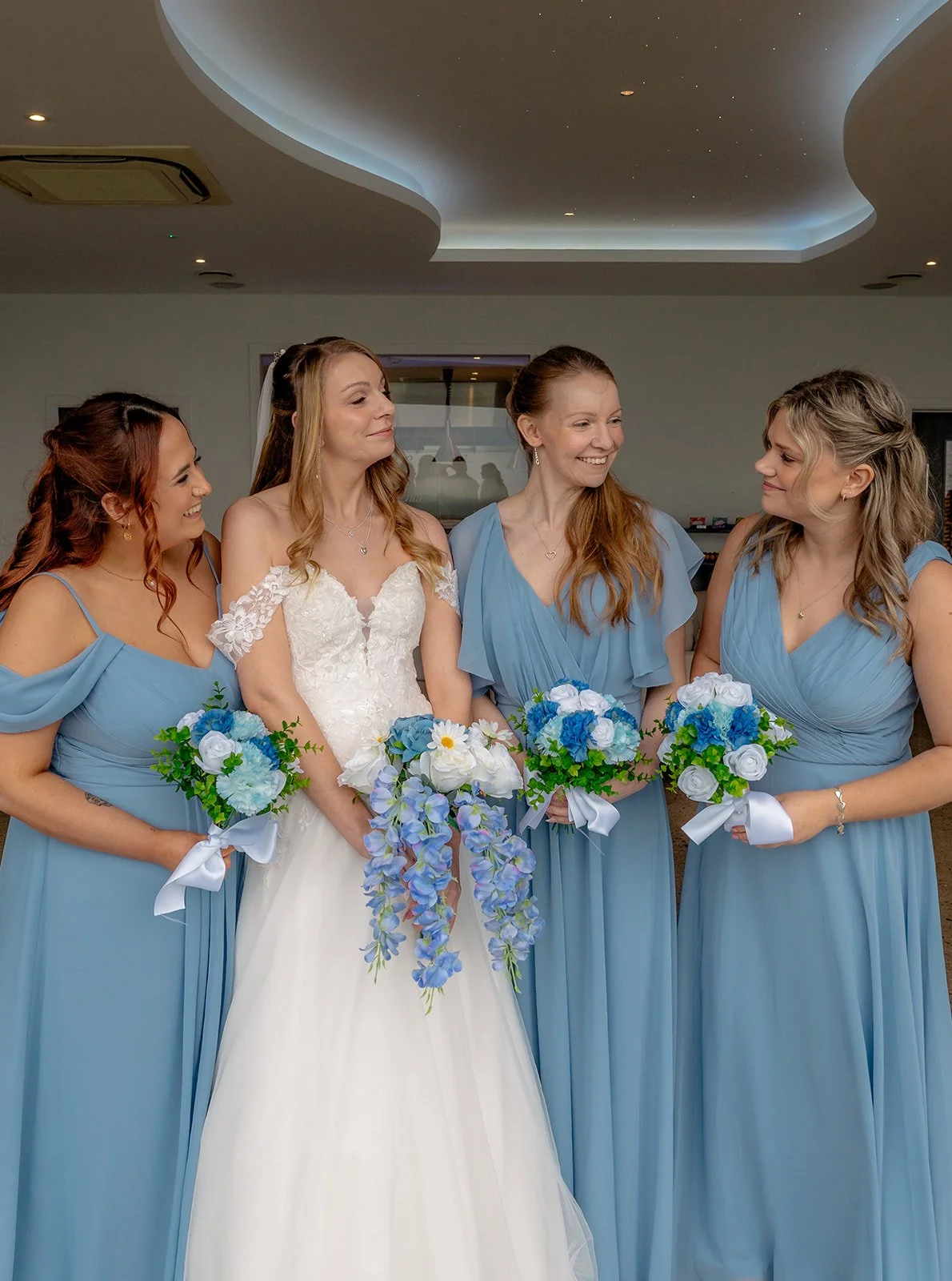 Bride Hannah stands with her bridesmaids in soft blue dresses during bridal party portraits at Sandy Cove Hotel, North Devon. The group smile at each other while holding blue and white bouquets in a relaxed moment.