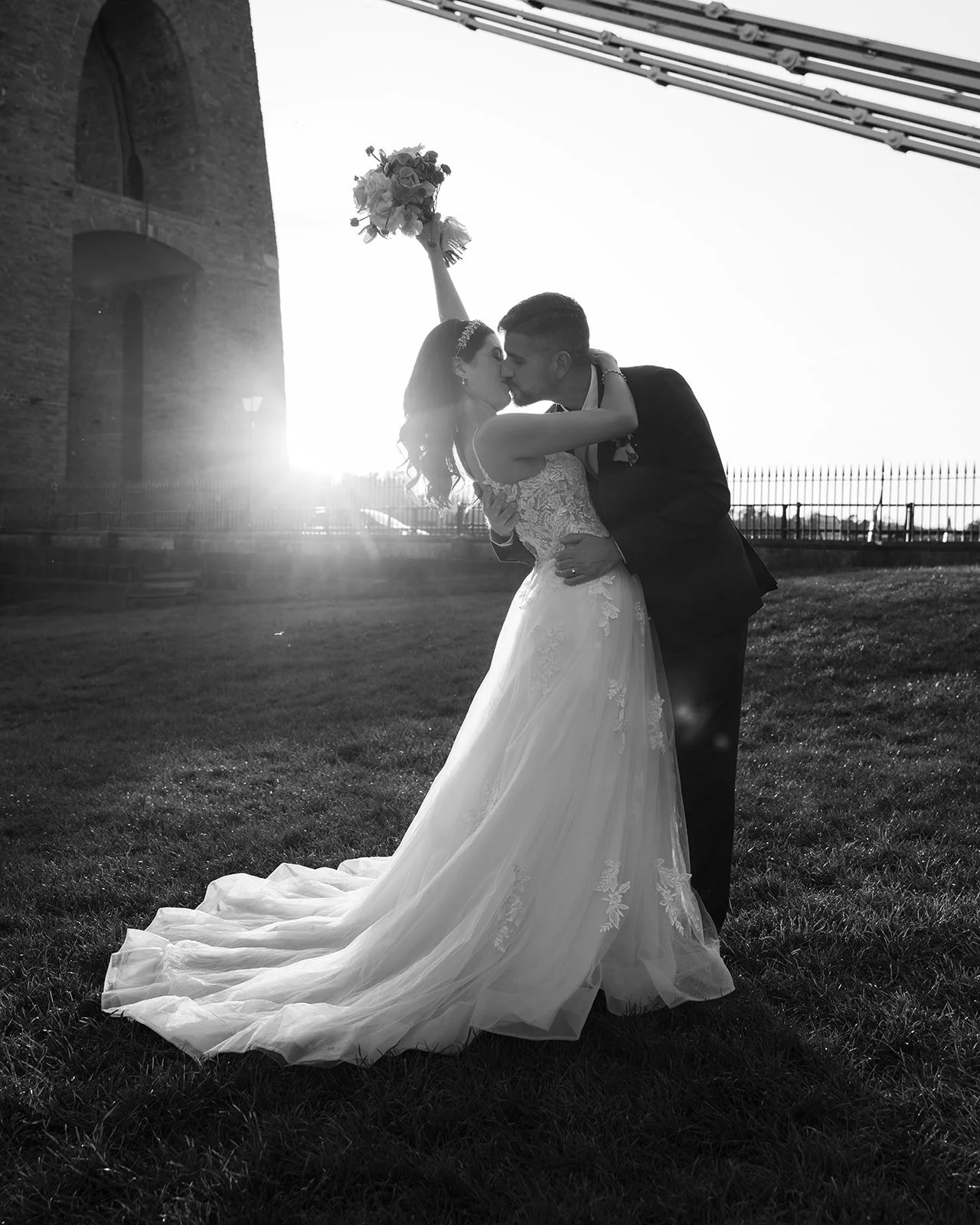 Black and white wedding portrait of Ruth and Carmelo kissing at sunset near Clifton Suspension Bridge in Bristol, UK, with Ruth raising her bouquet against the dramatic backlight.