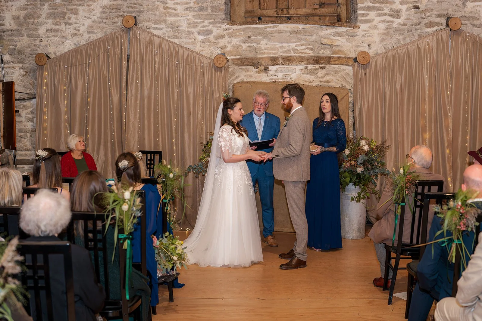 Maisie & Andrew holding hands during their indoor ceremony at Stockbridge Farm Barn in Dorset, surrounded by guests and rustic autumn wedding styling.