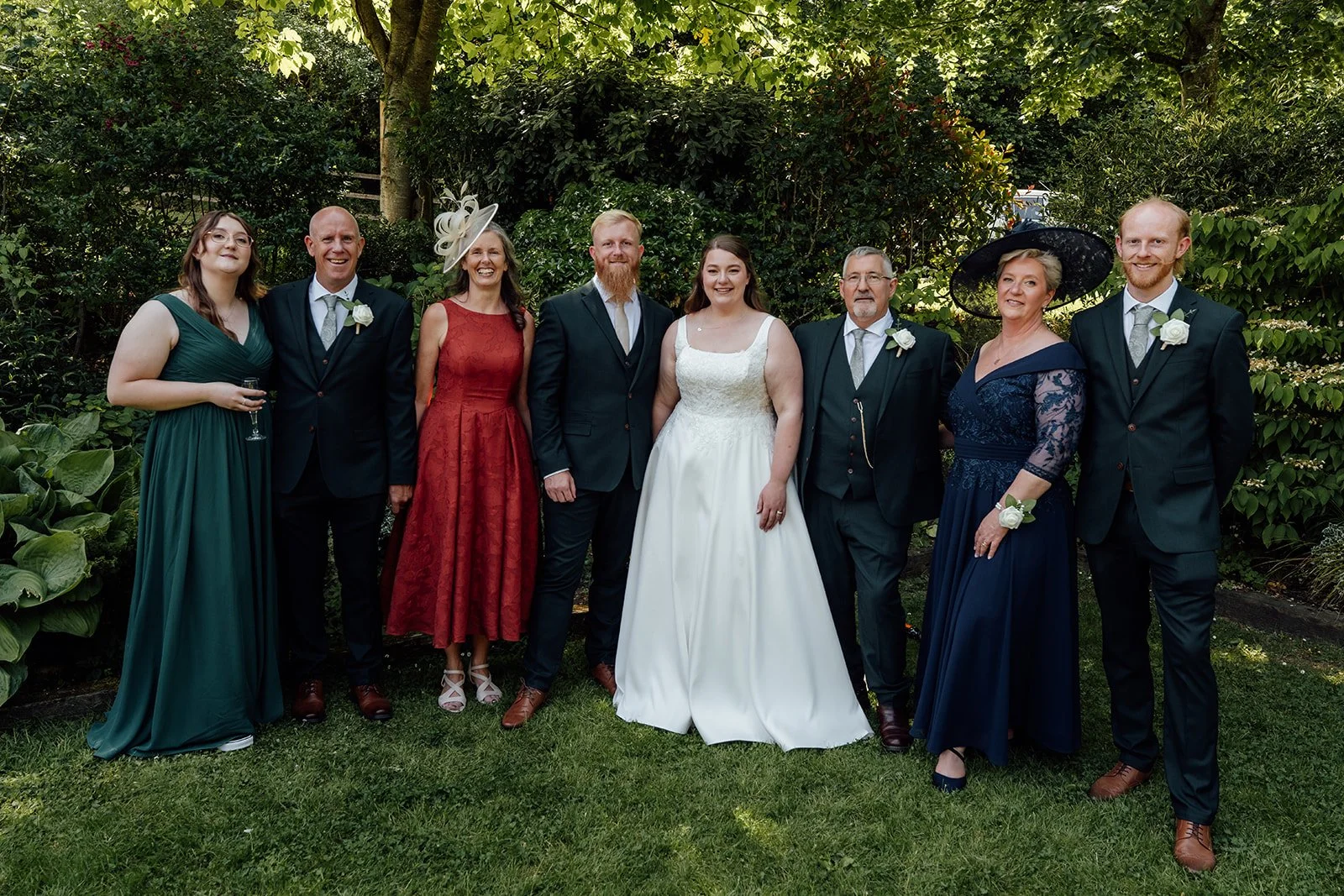 Family group portrait from Louise & Jon’s wedding at Wick Farm Bath in Somerset, UK, with the couple surrounded by loved ones in the garden, dark green suits, summer dresses, shaded greenery, and a classic countryside.