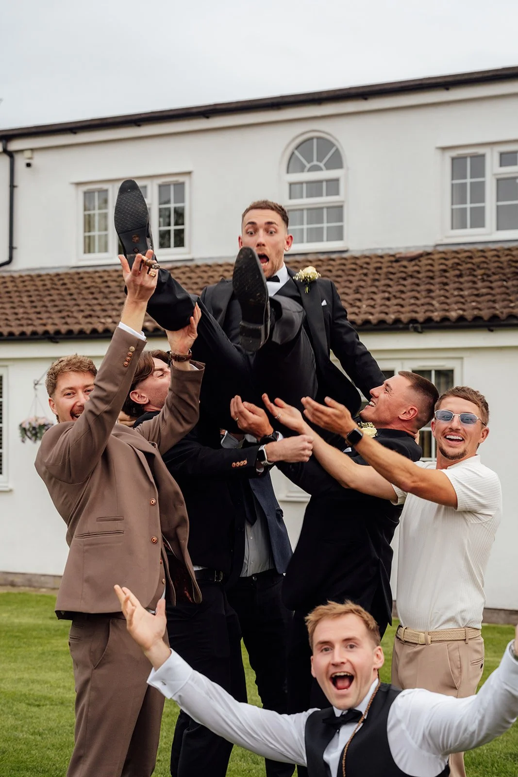 Jack is lifted into the air by friends during the wedding celebrations at Batch Country House in Somerset, UK, with guests laughing around him on the lawn as he reacts with surprise in his black tuxedo, photographed by Just James Weddings.
