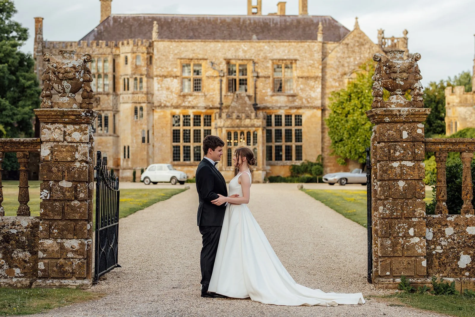 Rachel & Tom pose together on the driveway in front of historic Brympton House in Somerset, framed by the venue’s stone entrance gates, photographed by Just James Weddings.