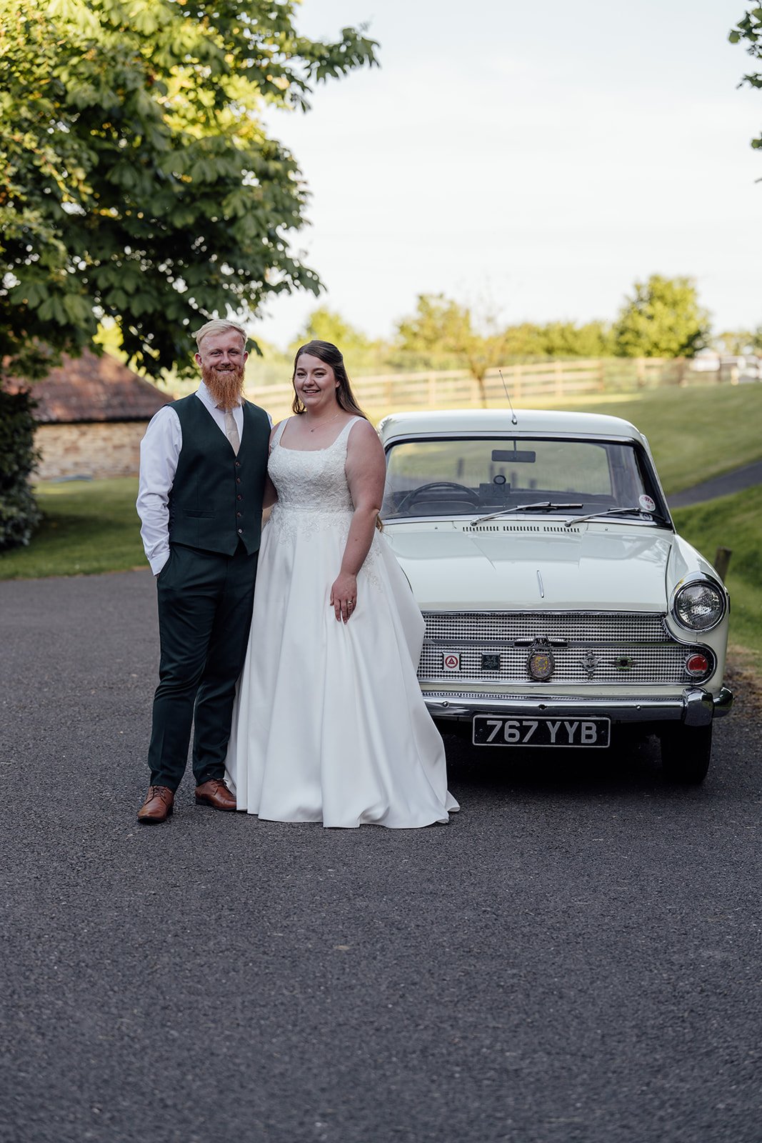 Classic couple portrait of Louise & Jon at Wick Farm Bath in Somerset, UK, standing beside a vintage cream wedding car on the lane with countryside views, soft evening light, white dress, green waistcoat, and relaxed newlywed styling together.