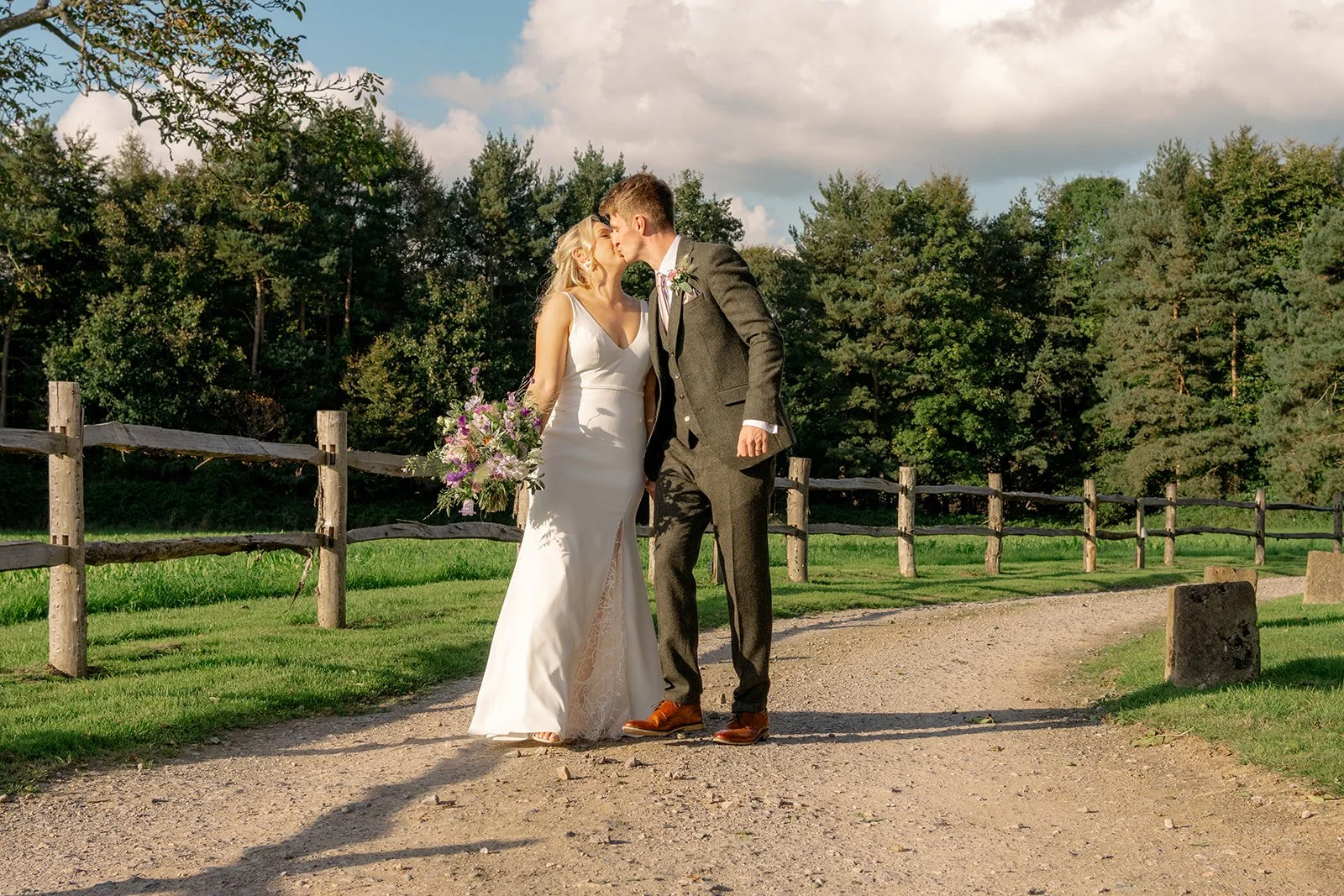 Abi & Josh kissing beside a wooden fence at The Cider Barn in Somerset, with countryside fields and woodland behind them, photographed by Just James Weddings for relaxed barn wedding portraits.