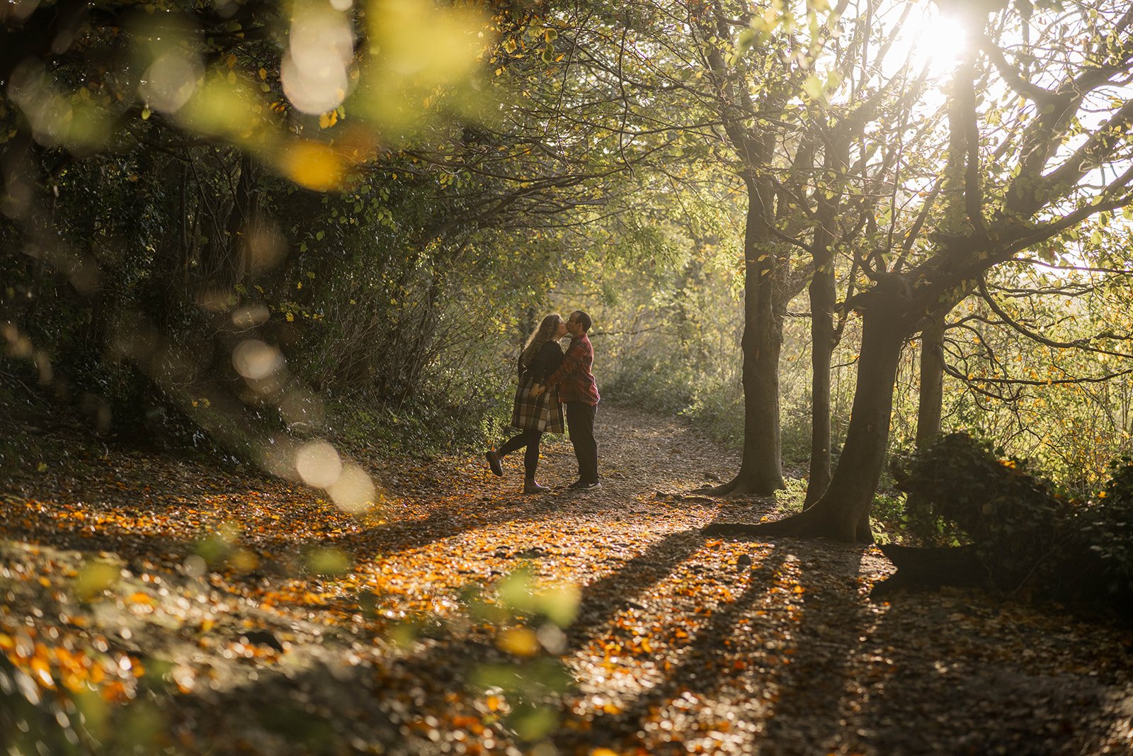 Kat & Ken share a kiss during their engagement shoot on a leaf-covered woodland path, framed by glowing autumn light, trees and soft foreground bokeh. Long shadows and golden tones create a warm, intimate and cinematic feel across the scene.