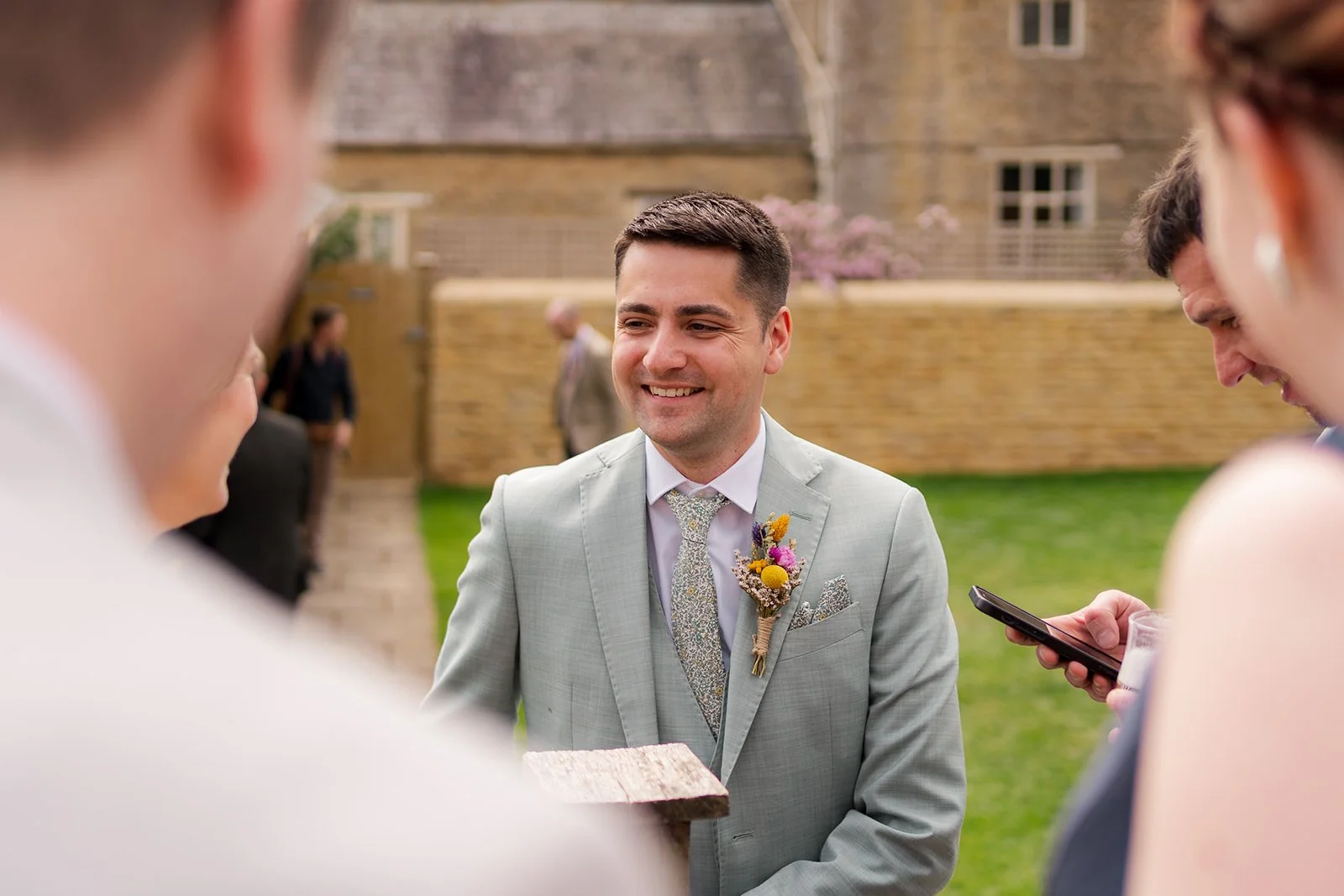 Sam smiling during the ceremony at The Great Barn at Aynho in the Cotswolds, surrounded by guests outdoors, photographed by Just James Weddings for candid wedding ceremony moments.