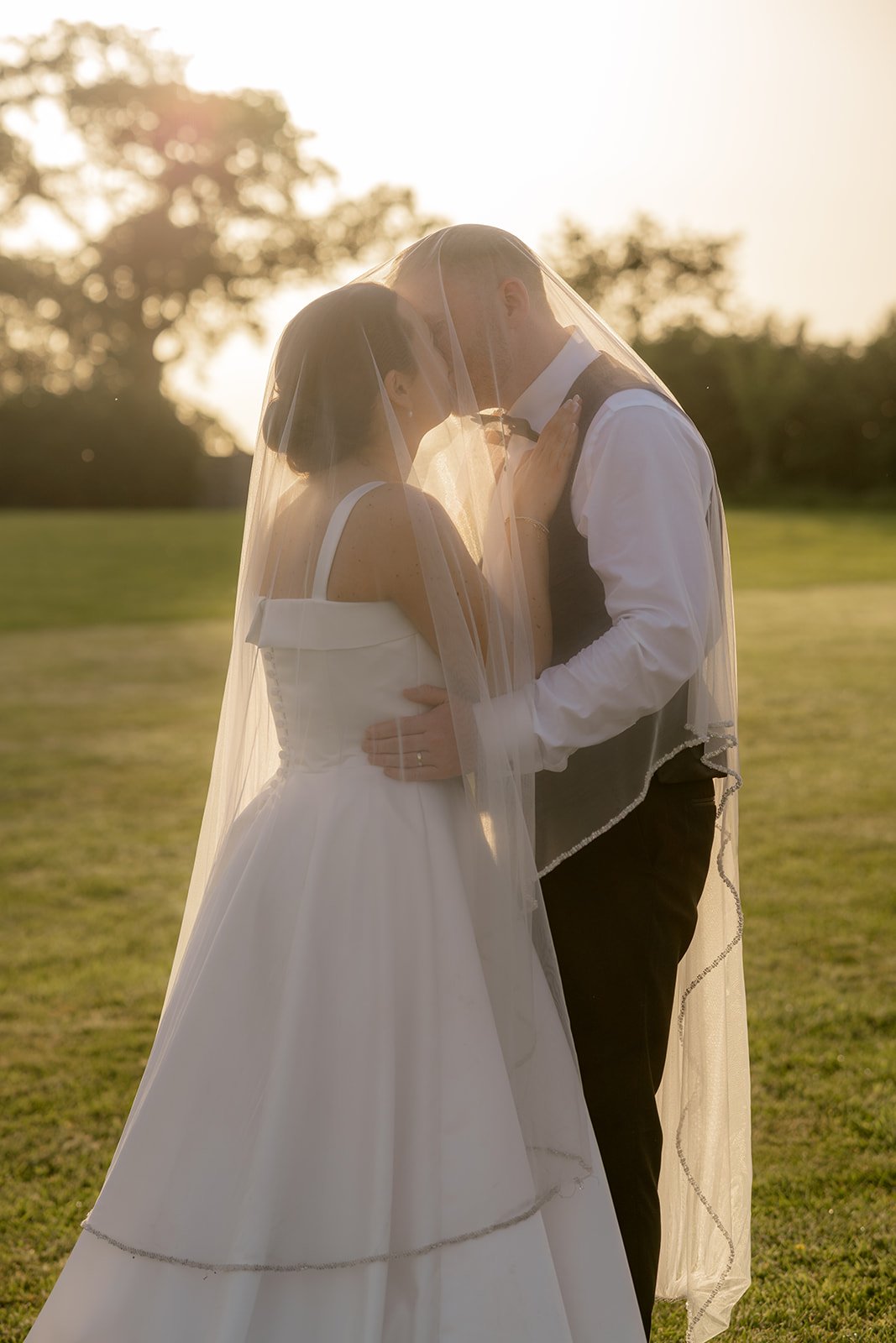 Golden hour portrait of Lydia & Kieran kissing beneath the bride's veil in a green field at Hencote Estate in Shropshire, UK, with soft backlight, countryside views and romantic outdoor wedding photography from their summer celebration day.