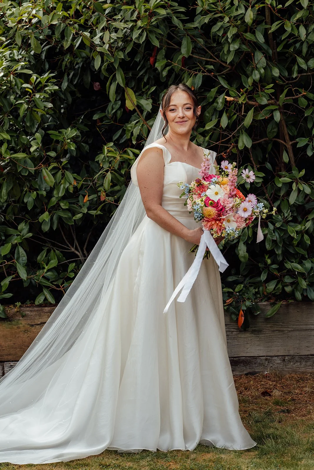 Harriet poses with her colourful pink, orange and blue bouquet against leafy greenery at Dovecote Events in Oxfordshire, photographed by Just James Weddings.