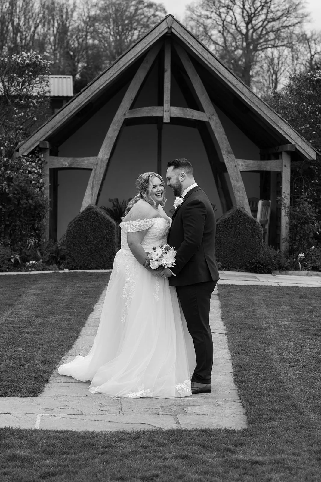 Black and white portrait of Siobhan & Nicholas standing together on the garden path at Kingscote Barn in the Cotswolds, UK, smiling outside the rustic barn wedding venue, a timeless countryside wedding portrait with bouquet, lawn and pavilion.