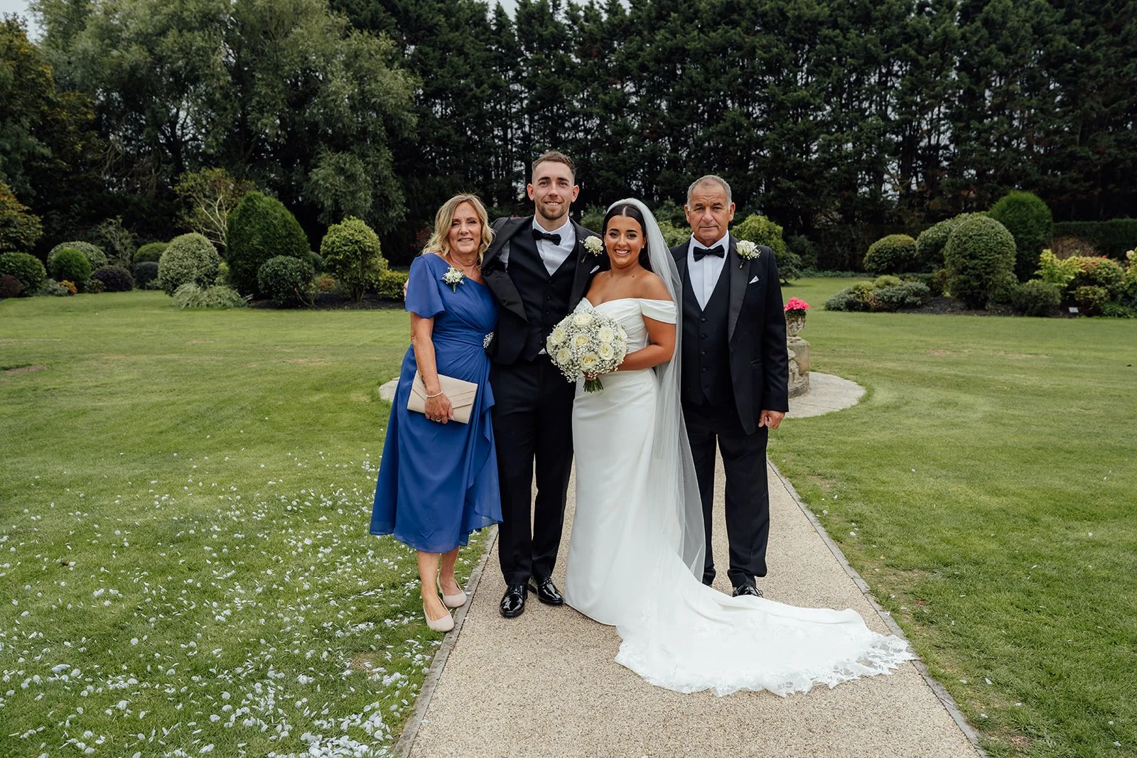 Nisha and Jack stand with two family members for a formal wedding portrait on the gardens at Batch Country House in Somerset, UK, with Nisha holding her white rose bouquet and Jack in his black tuxedo, photographed by Just James Weddings.