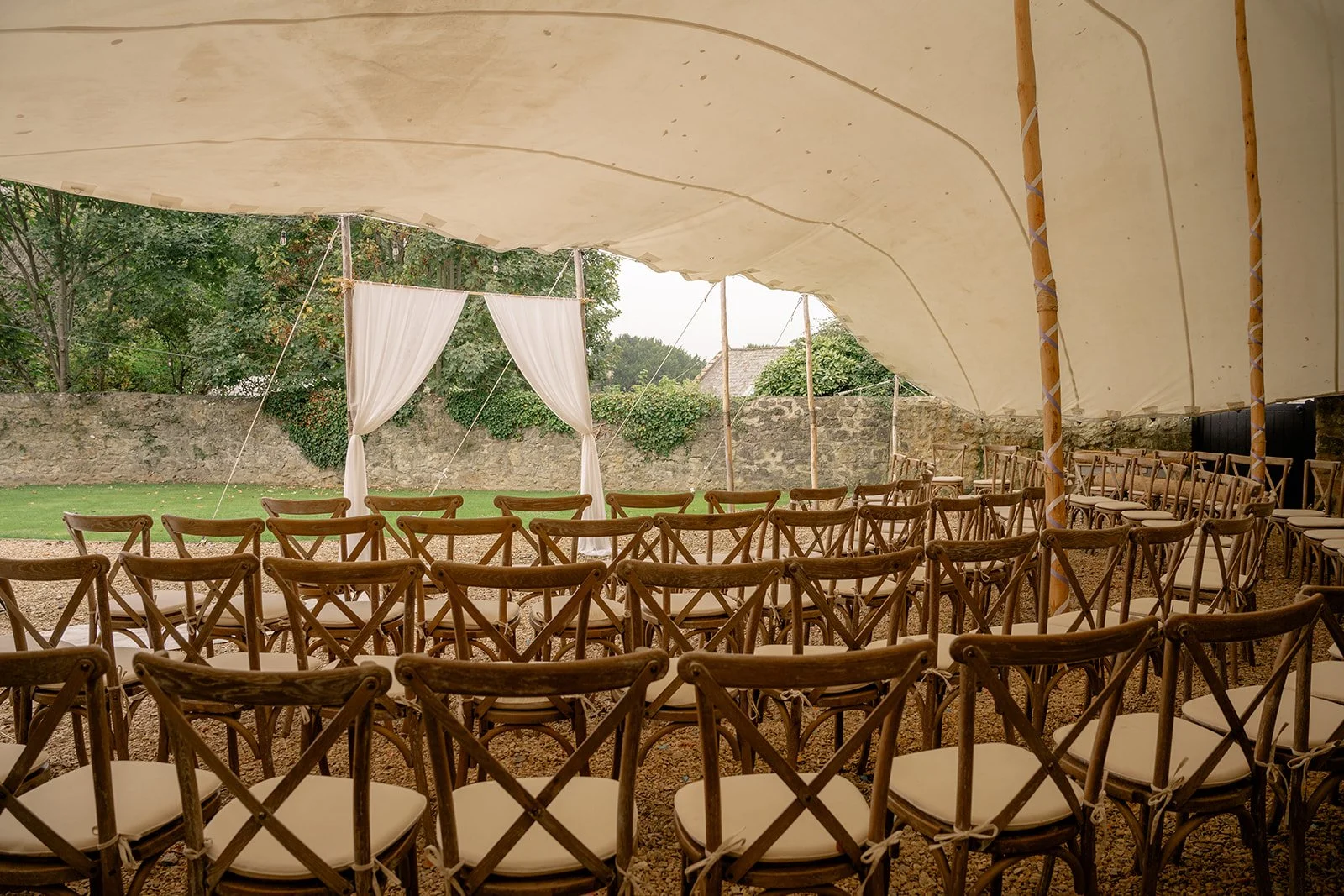 Outdoor ceremony setup under a canvas marquee at The Cider Barn in Somerset for Abi & Josh’s wedding, with wooden chairs, white drapes and stone garden walls, photographed by Just James Weddings for rustic countryside wedding ceremony.