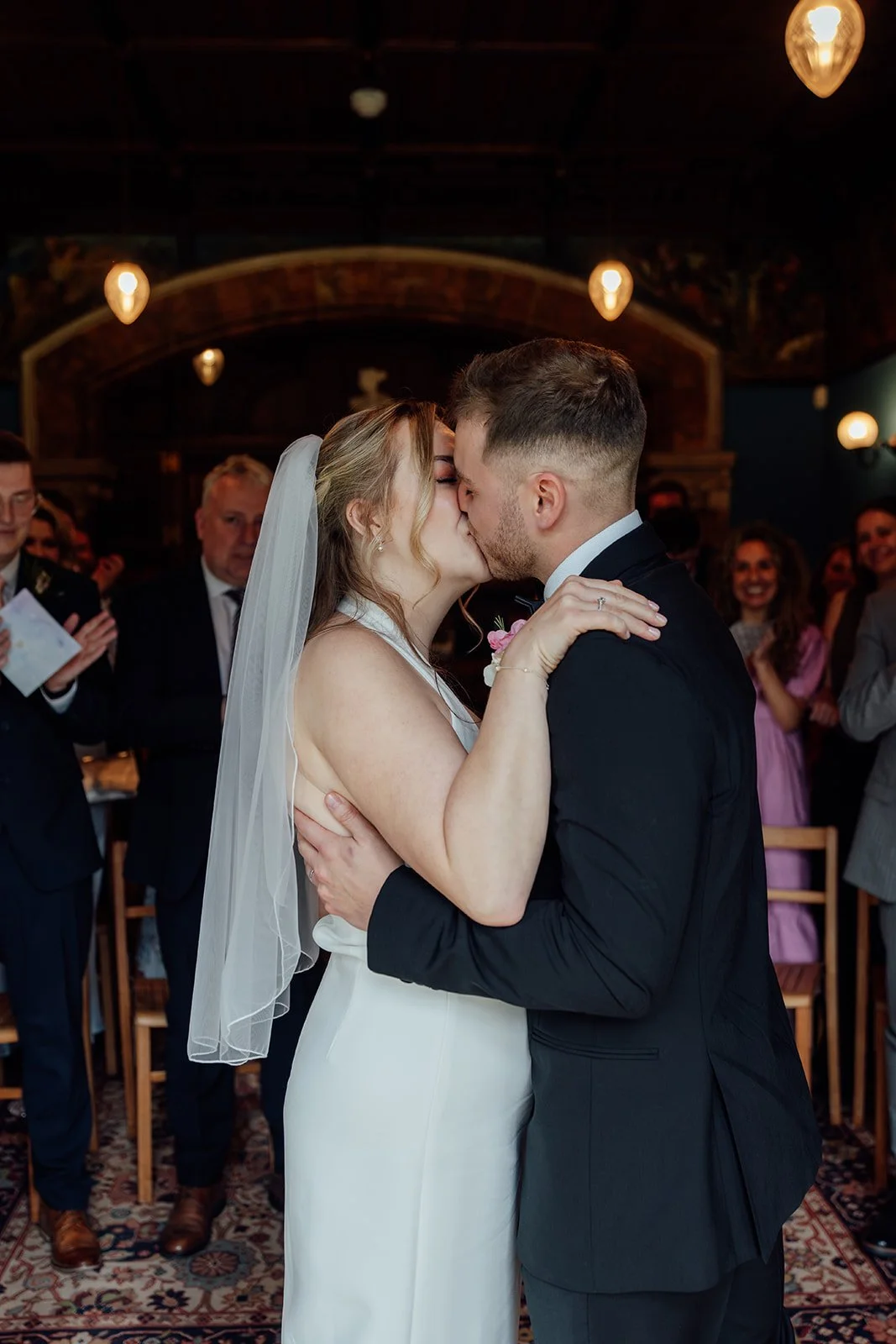 Bryony & Max share their first kiss during their wedding ceremony at Insole Court in Cardiff, surrounded by smiling guests, warm pendant lights, rich interiors and soft applause after their intimate vows in the historic ceremony room space.