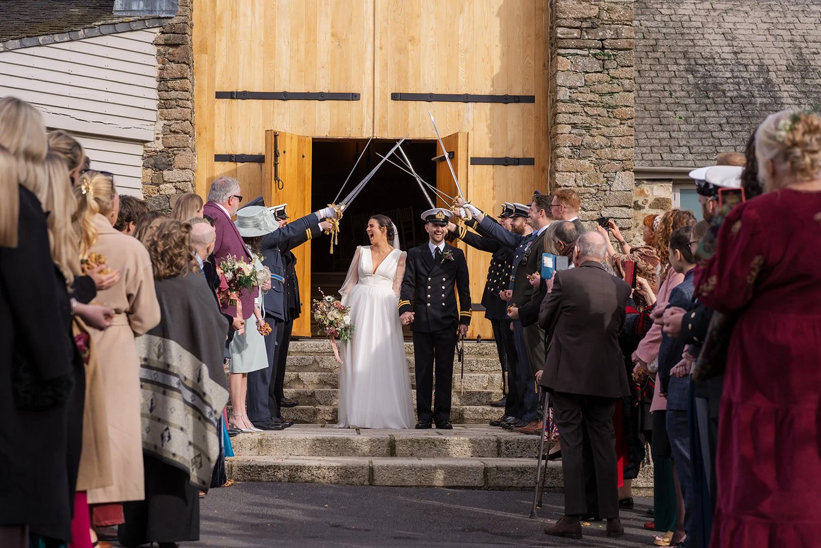 Megan & Luca walk hand in hand beneath raised naval swords at The Great Barn Devon as guests cheer and smile around them. The wooden barn doors and stone walls frame this joyful Devon wedding celebration.
