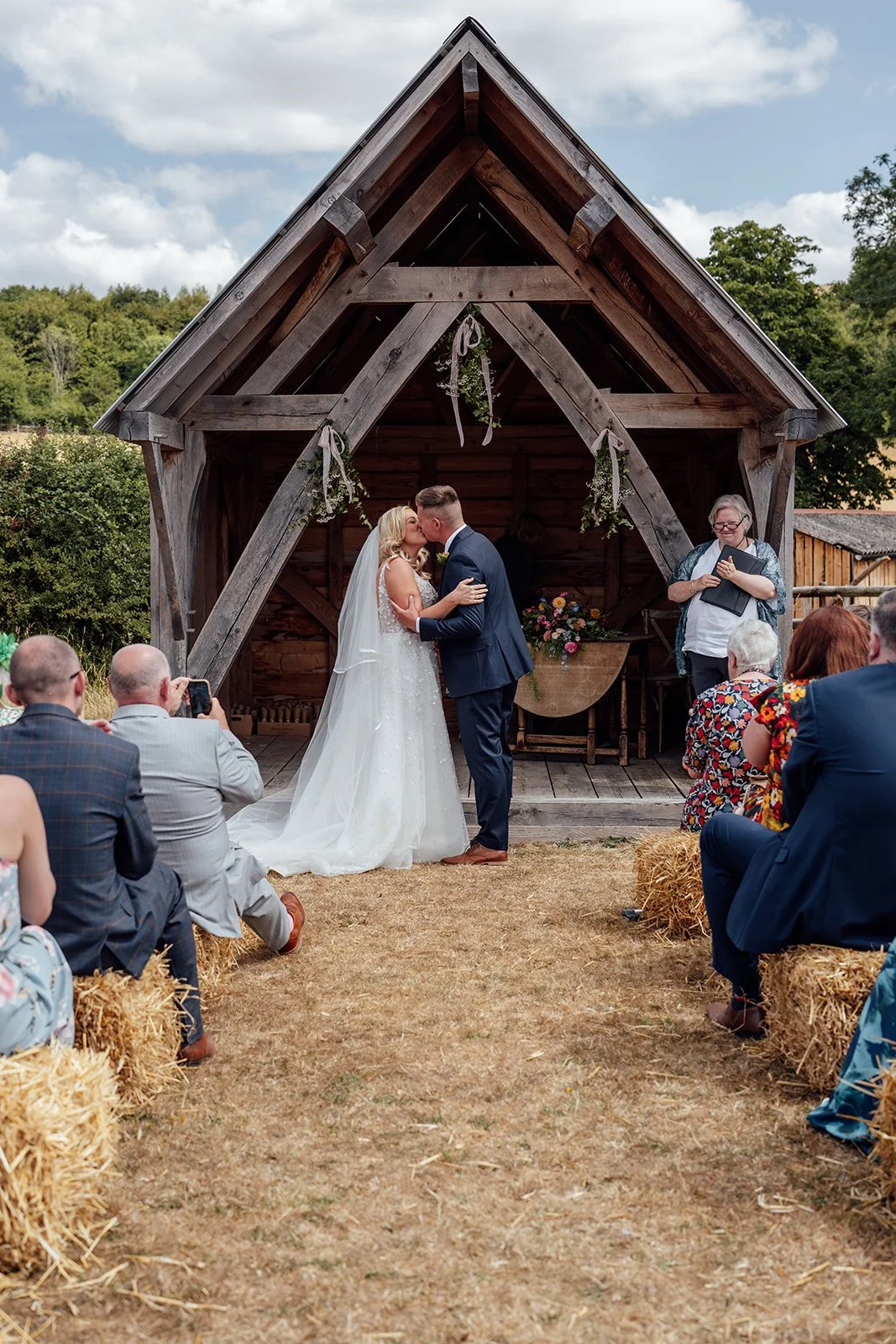 Elise and Tom sharing their first kiss during their outdoor wedding ceremony beneath the timber structure at Bowerchalke Barn in Wiltshire, photographed by Just James Weddings.