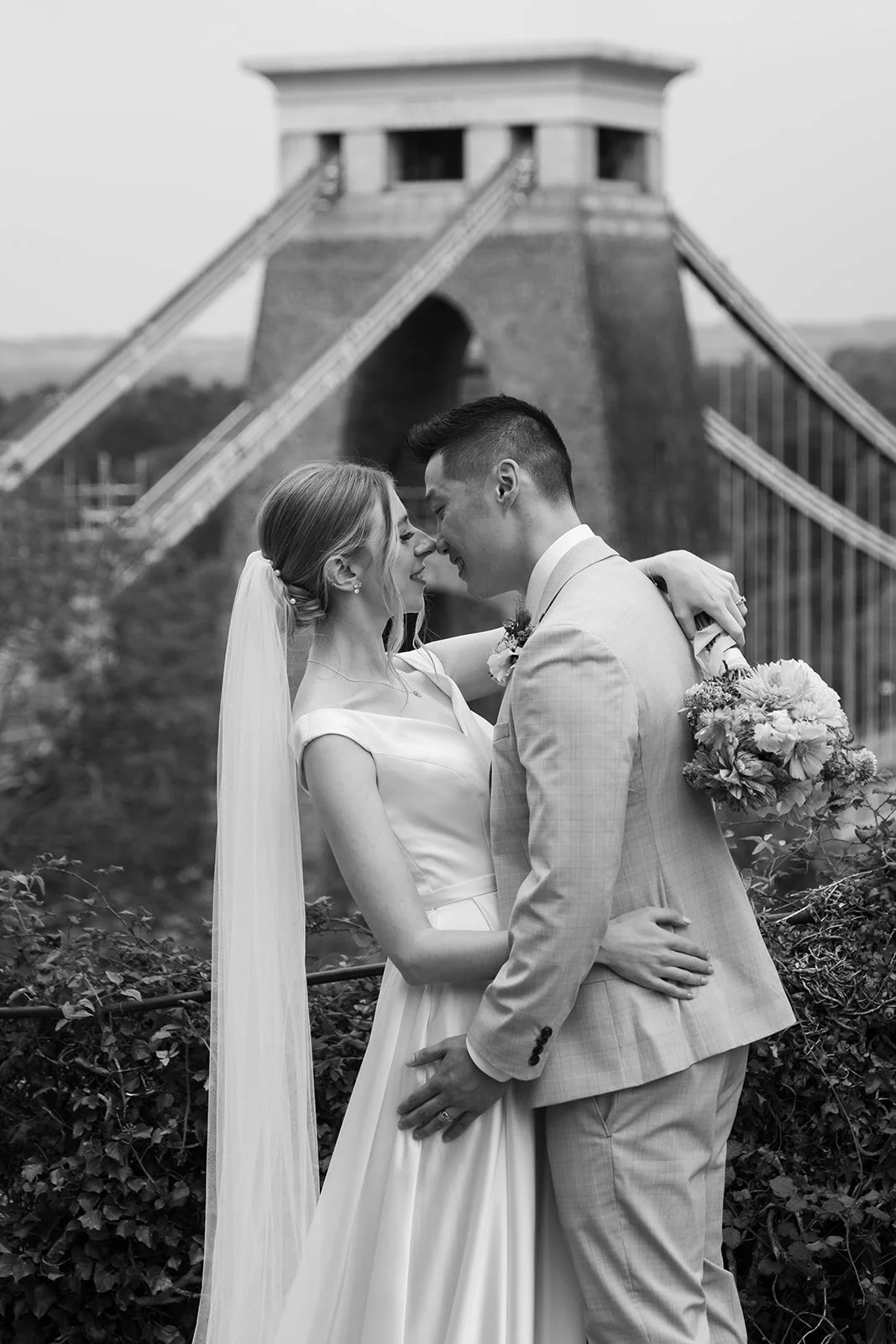 Black and white portrait of Brian & Josie standing together at Clifton Observatory in Somerset, with Clifton Suspension Bridge behind them, photographed by Just James Weddings.