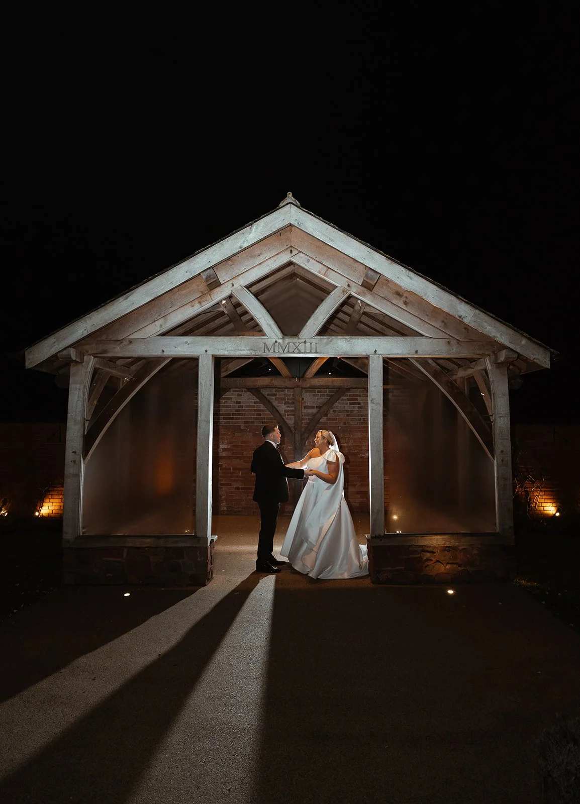 Jessie & Jay stand beneath the timber pavilion at Upton Barn & Walled Garden in Devon, UK, lit softly against the night sky. Long shadows and warm lighting frame a cinematic evening portrait, capturing a quiet, romantic moment together.