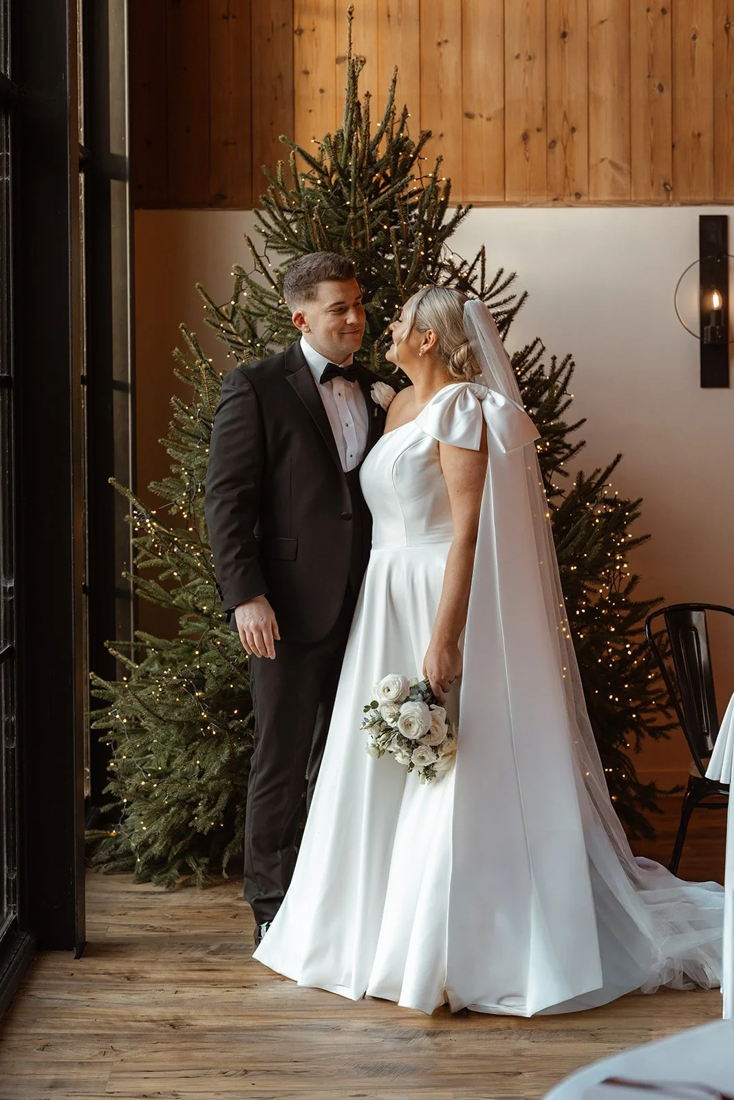 Jessie & Jay stand together inside Upton Barn & Walled Garden in Devon, UK, posing in front of a softly lit Christmas tree. Warm wooden textures and fairy lights frame a cosy, festive portrait from their winter wedding celebration.
