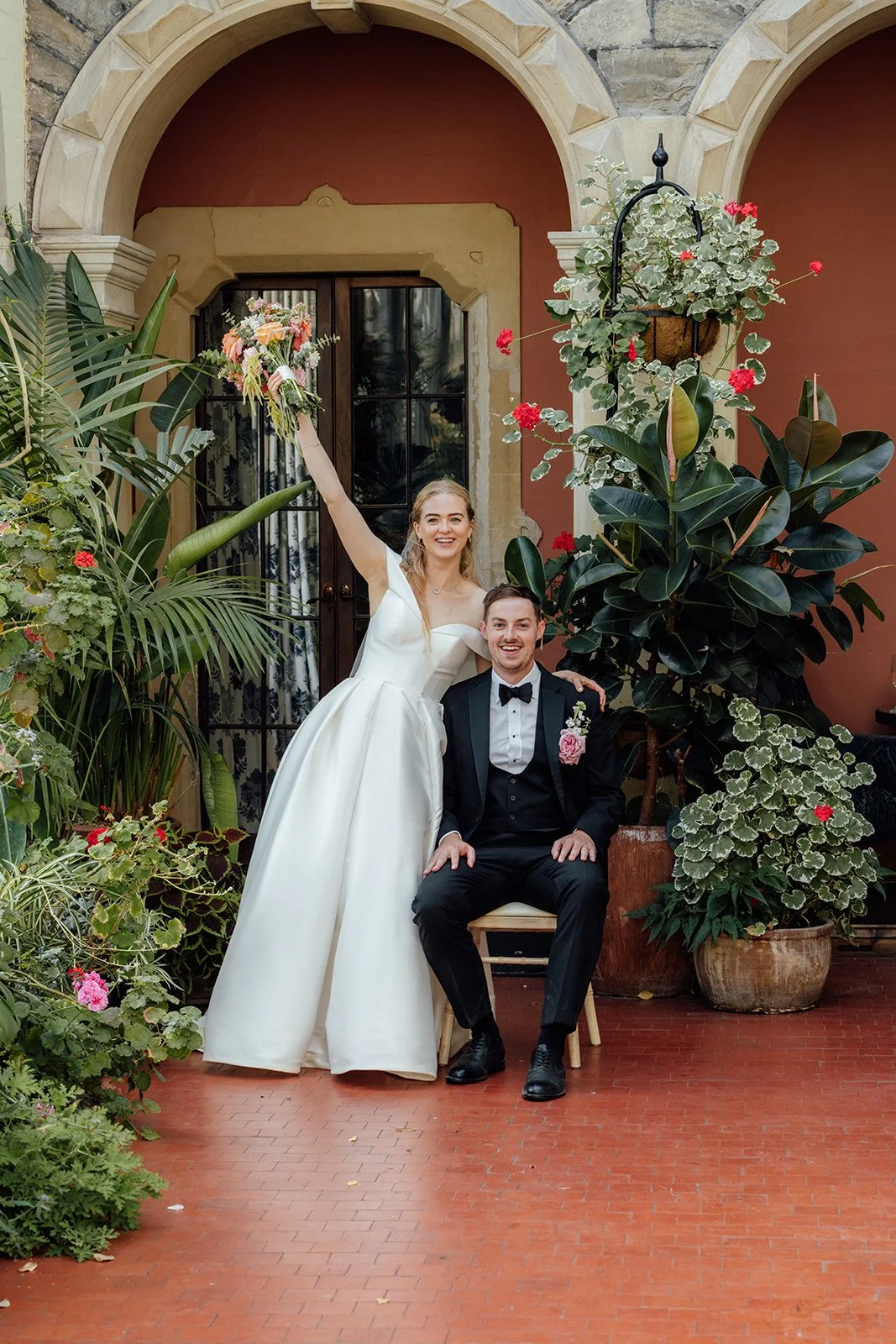 Caitlin raising her colourful bridal bouquet beside Kevin in a fun wedding portrait among the lush plants and stone arches at Clevedon Hall in Somerset.