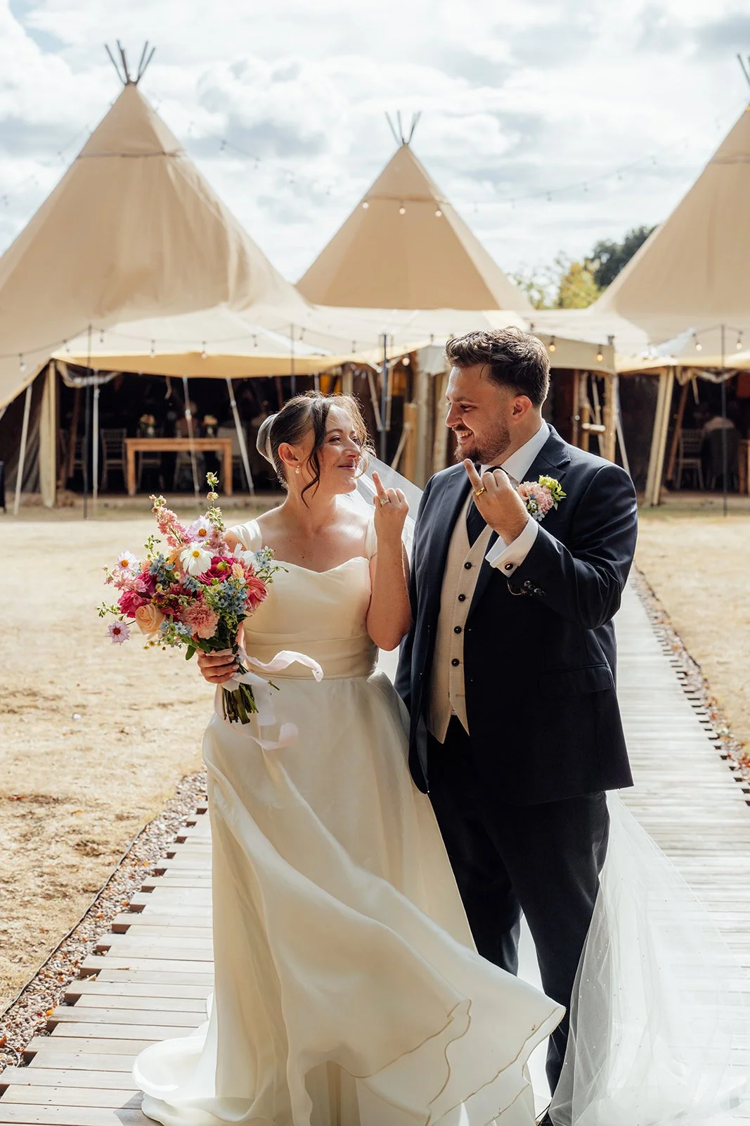 Harriet & Simon smile together outside the tipi reception space, with Harriet holding her colourful bouquet at Dovecote Events in Oxfordshire, photographed by Just James Weddings.