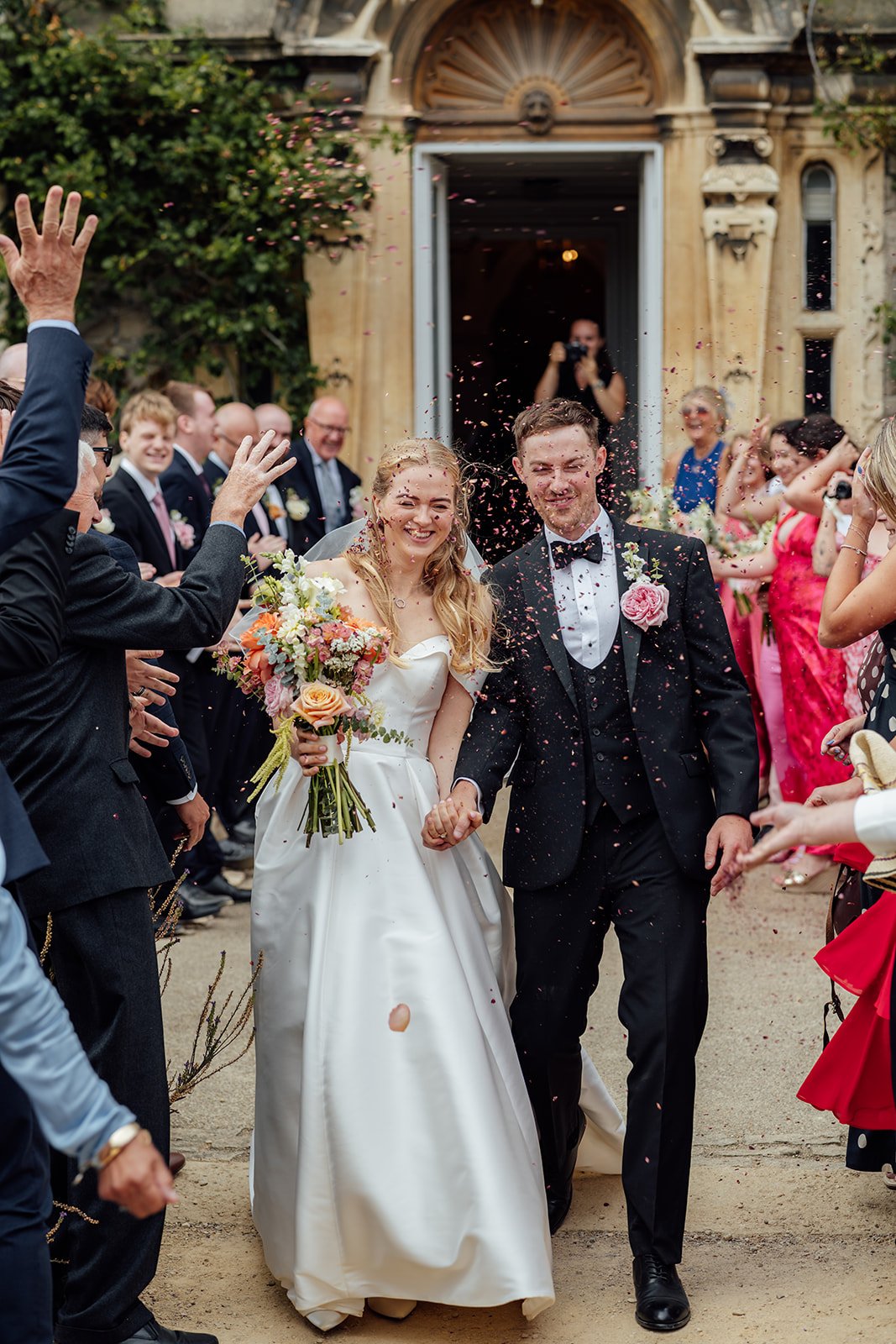 Caitlin & Kevin walking through confetti after their ceremony at Clevedon Hall in Somerset, surrounded by cheering guests, photographed by Just James Weddings in a joyful documentary style.